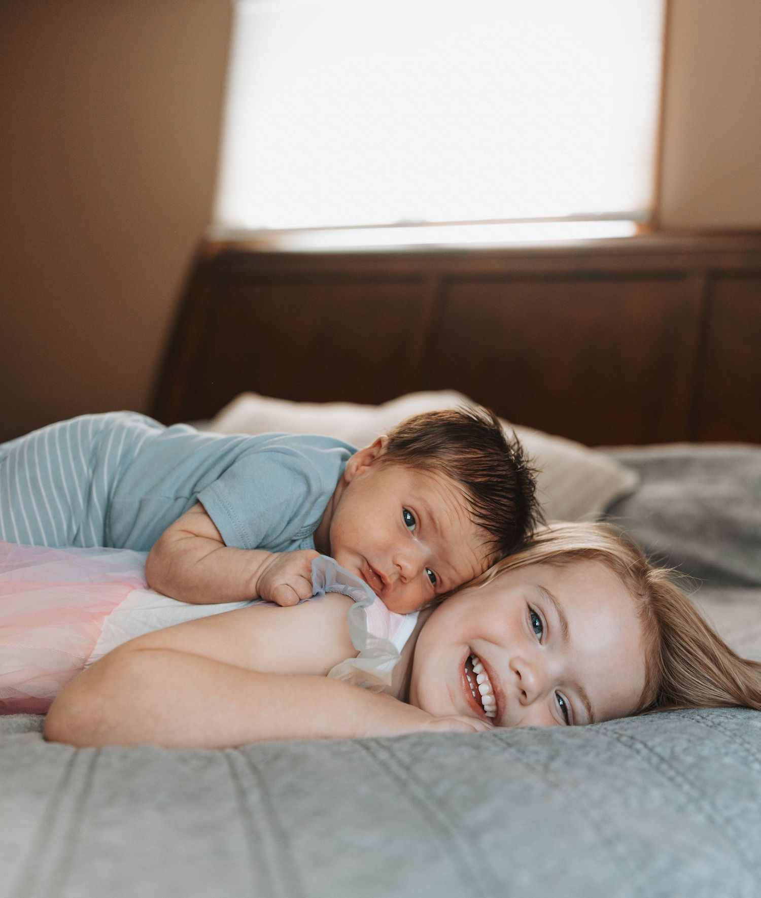 Two young children smiling and lying together on a bed, sharing a joyful sibling moment indoors.