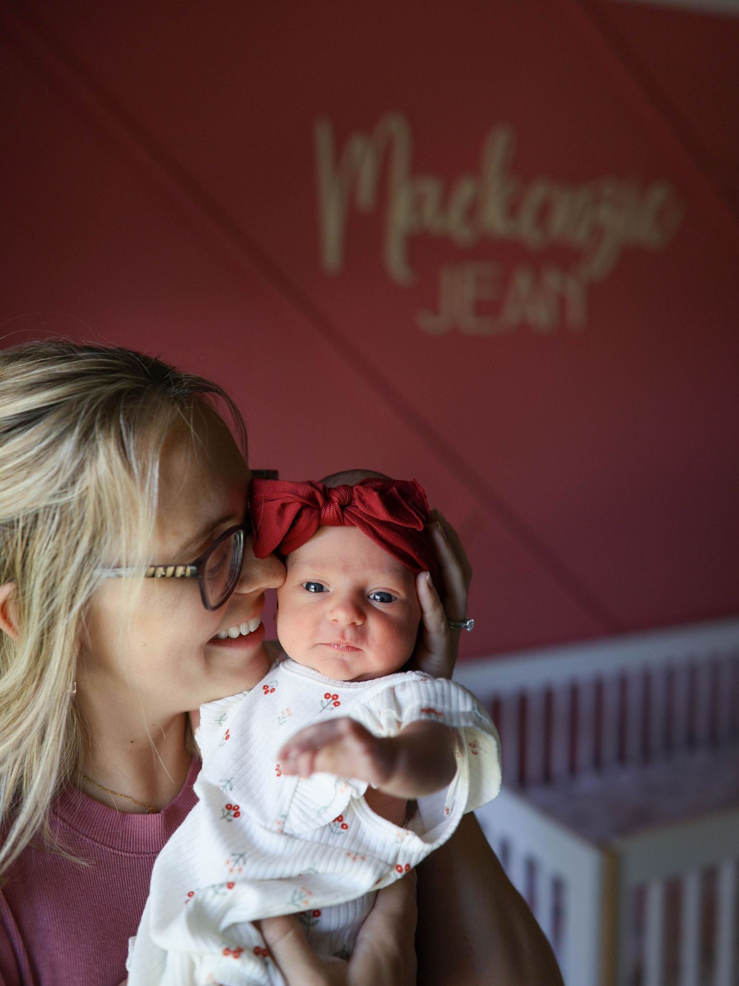 Smiling blonde mother holding newborn baby girl in red bow headband in nursery with name Mackenzie Jean on wall.