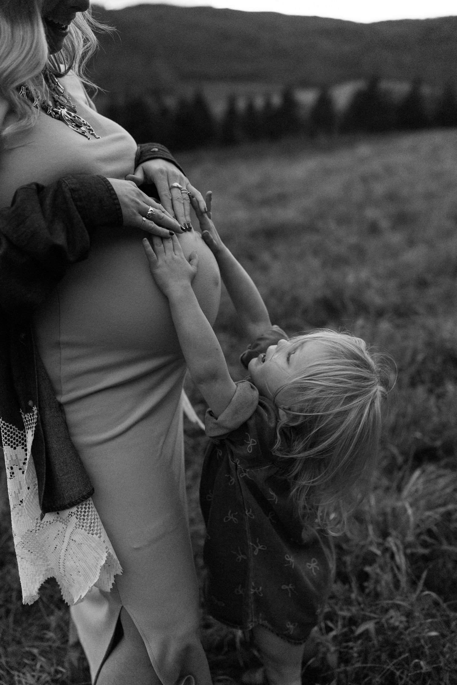 Black and white portrait showing tender embrace in a field with wind-blown hair and dramatic lighting.