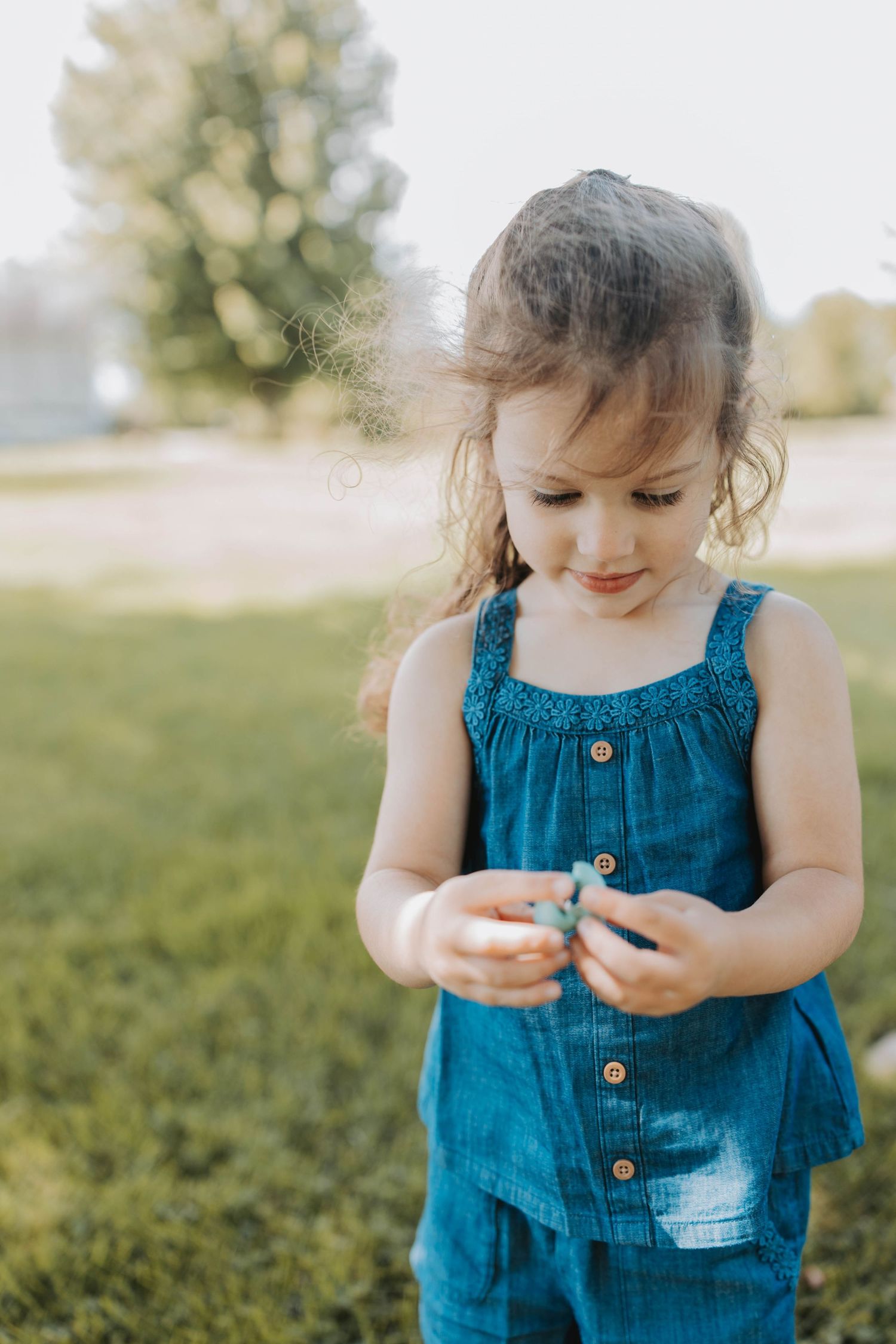 Young girl in blue denim romper looking down at something in her hands while standing outdoors in green grass.