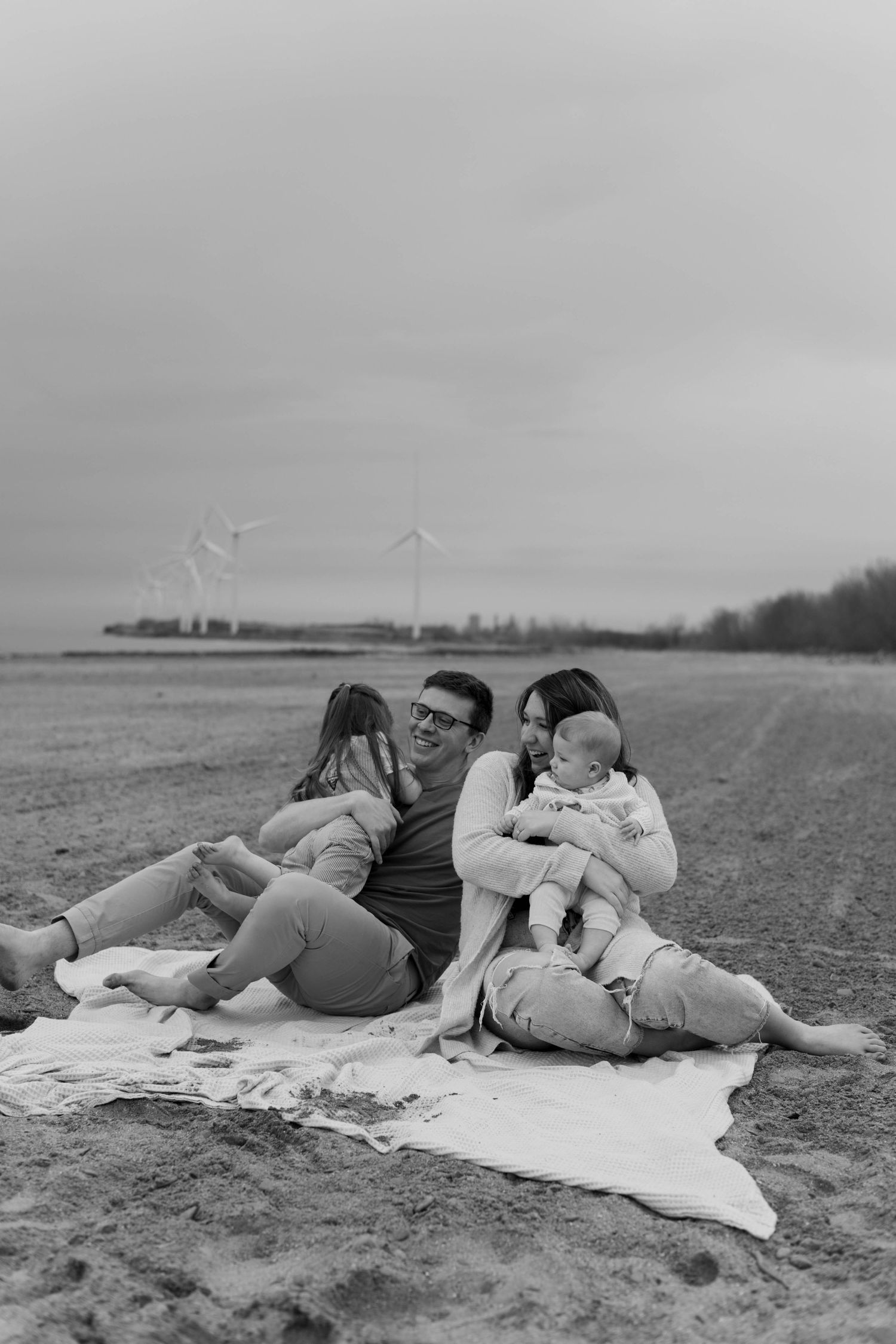 Black and white photo of a family relaxing on a blanket at the beach with wind turbines in the background.