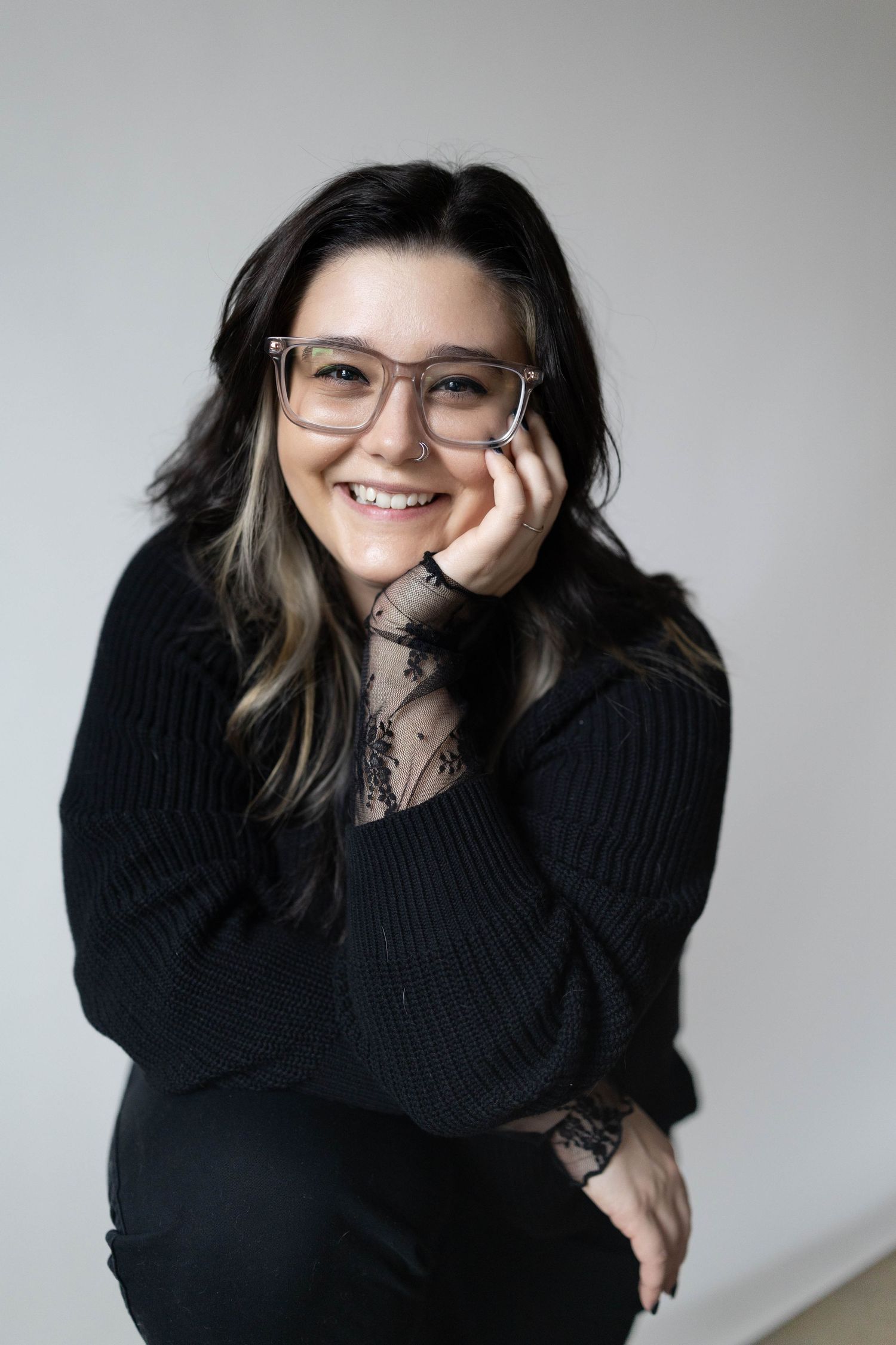 Person wearing glasses and black sweater sitting against white wall with arms resting on knees in casual portrait pose.