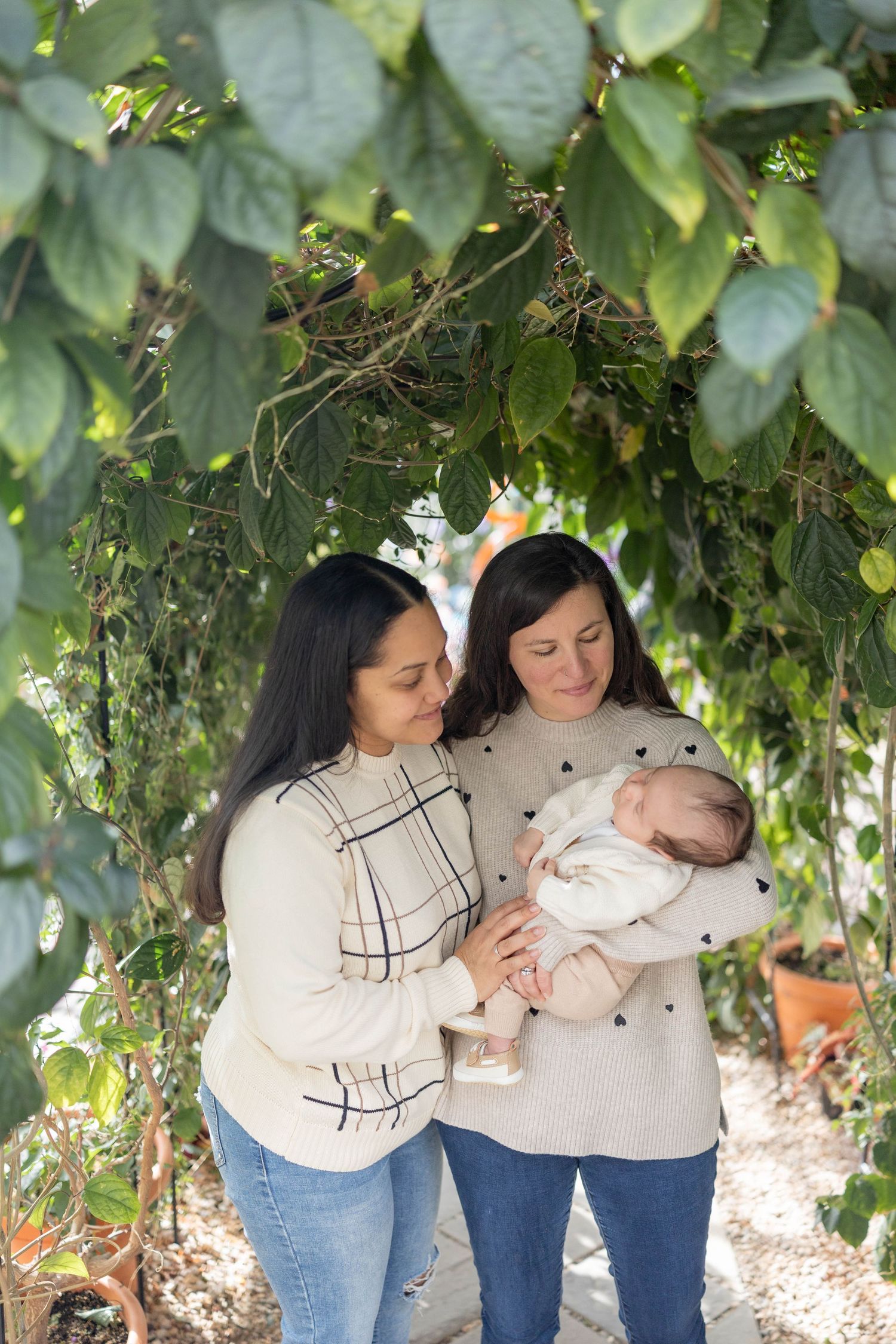 Two people in cream sweaters share a tender moment with a baby underneath lush green foliage.
