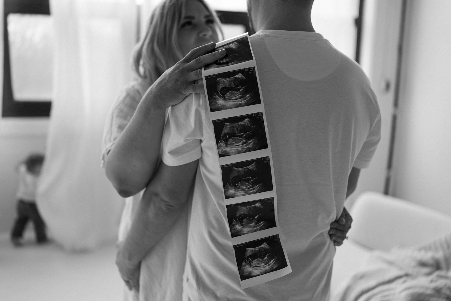 Black and white photo of couple holding ultrasound strip of sonogram images while embracing in a room.
