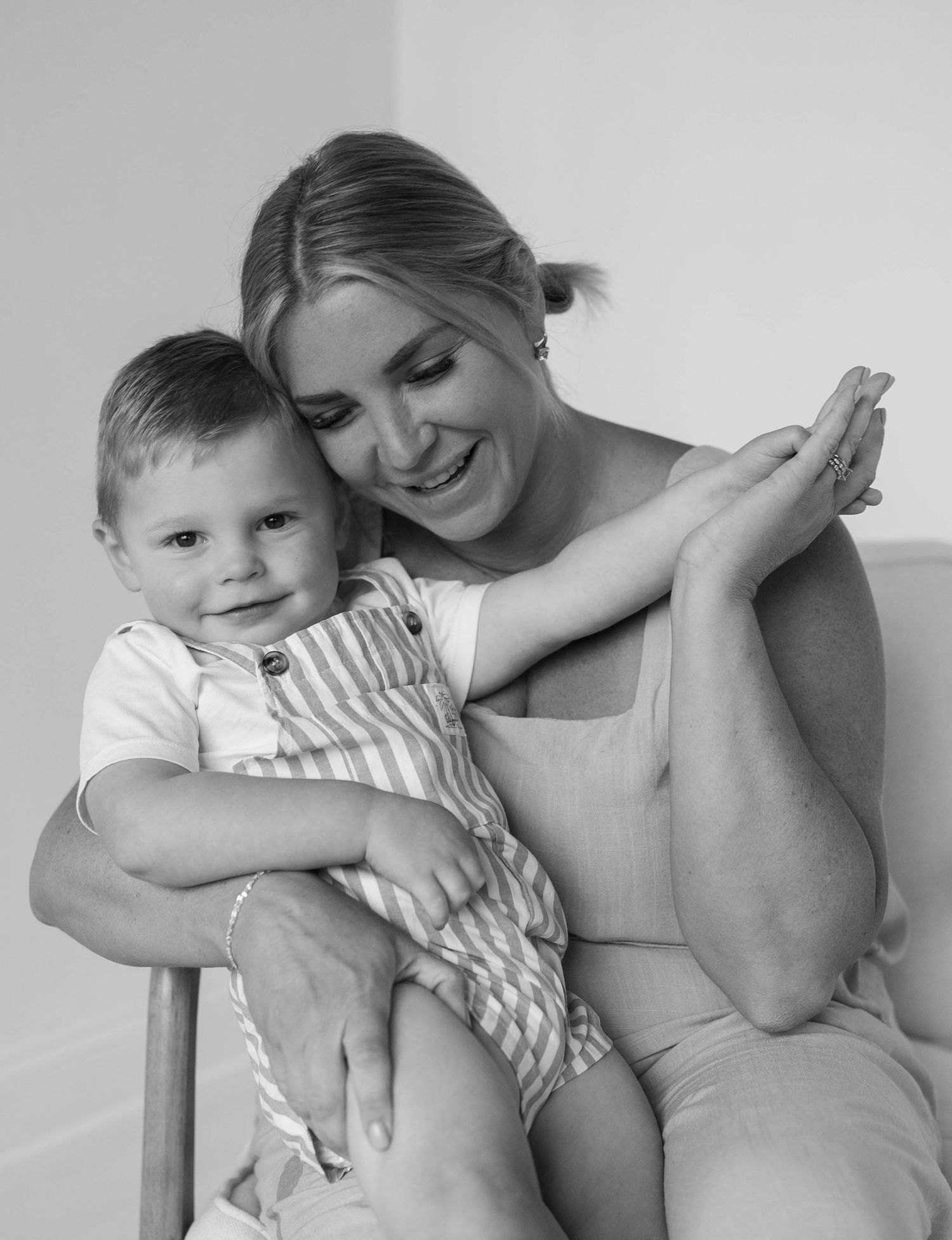 Black and white photo of a smiling woman sitting on a chair, lovingly holding a happy toddler boy in striped overalls.