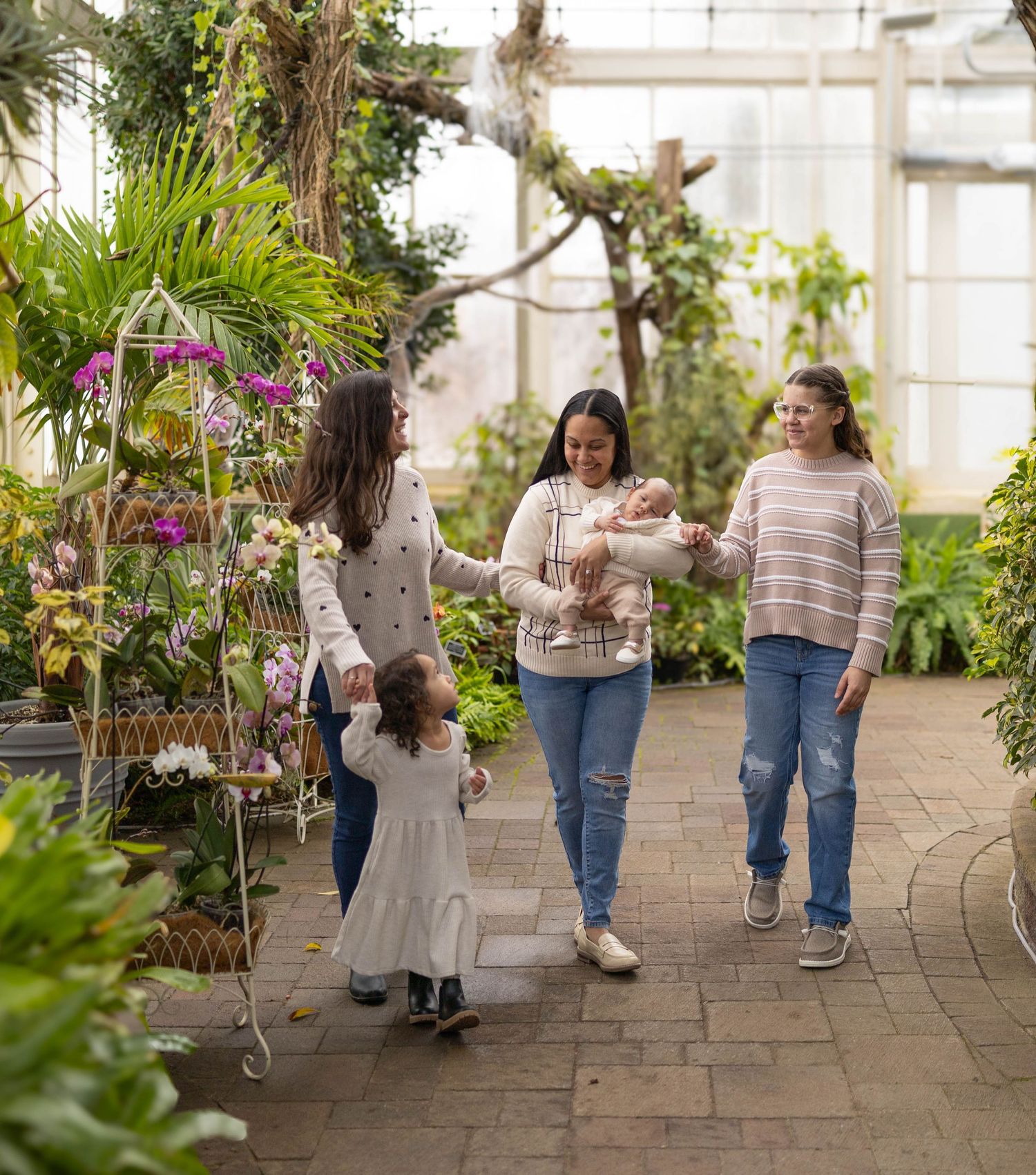 People walking together through a lush greenhouse filled with hanging plants and purple orchids on stone pathways.