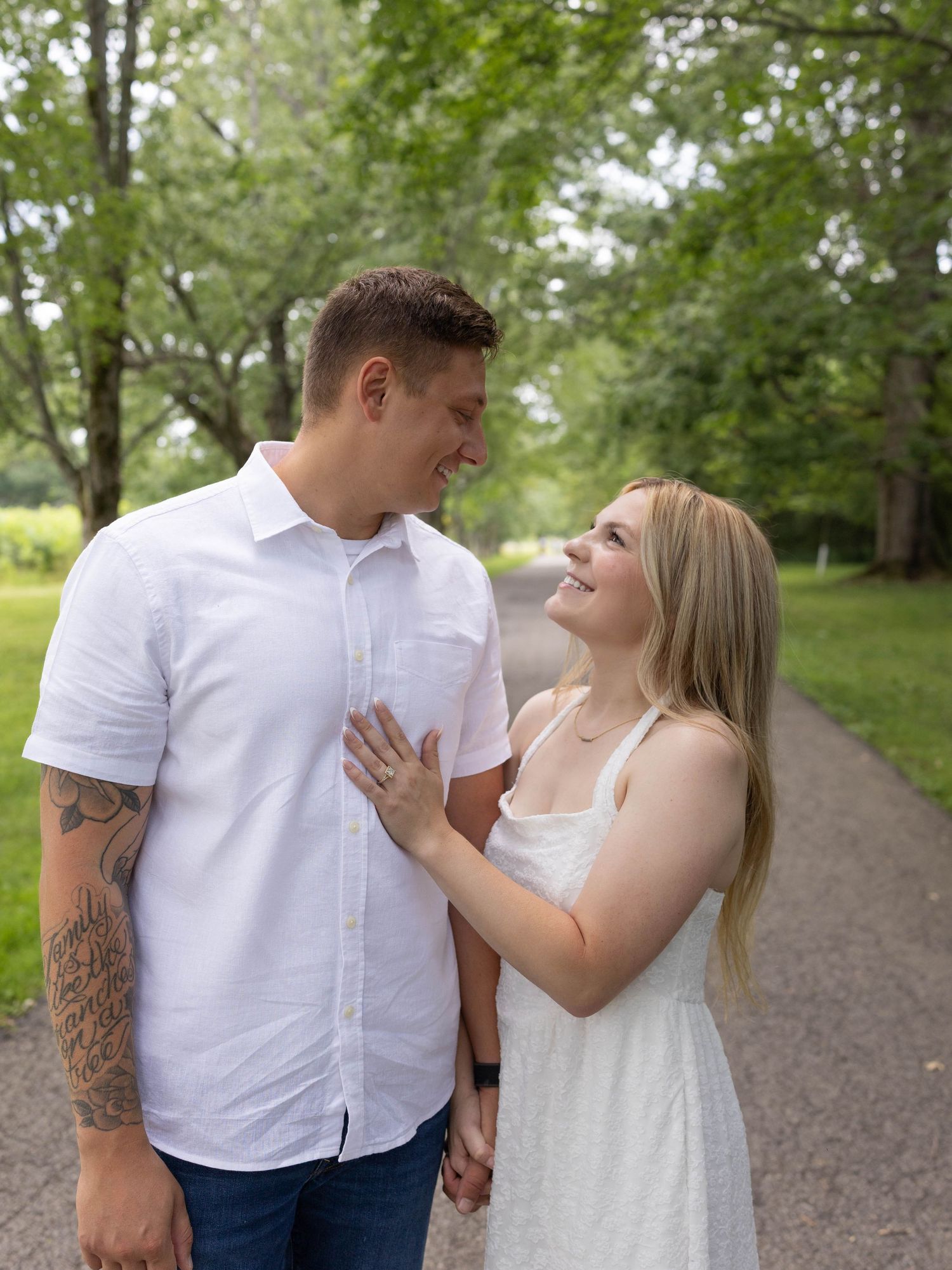 Couple smiling at each other on a tree-lined path, man with tattoos in white shirt, woman in white dress.