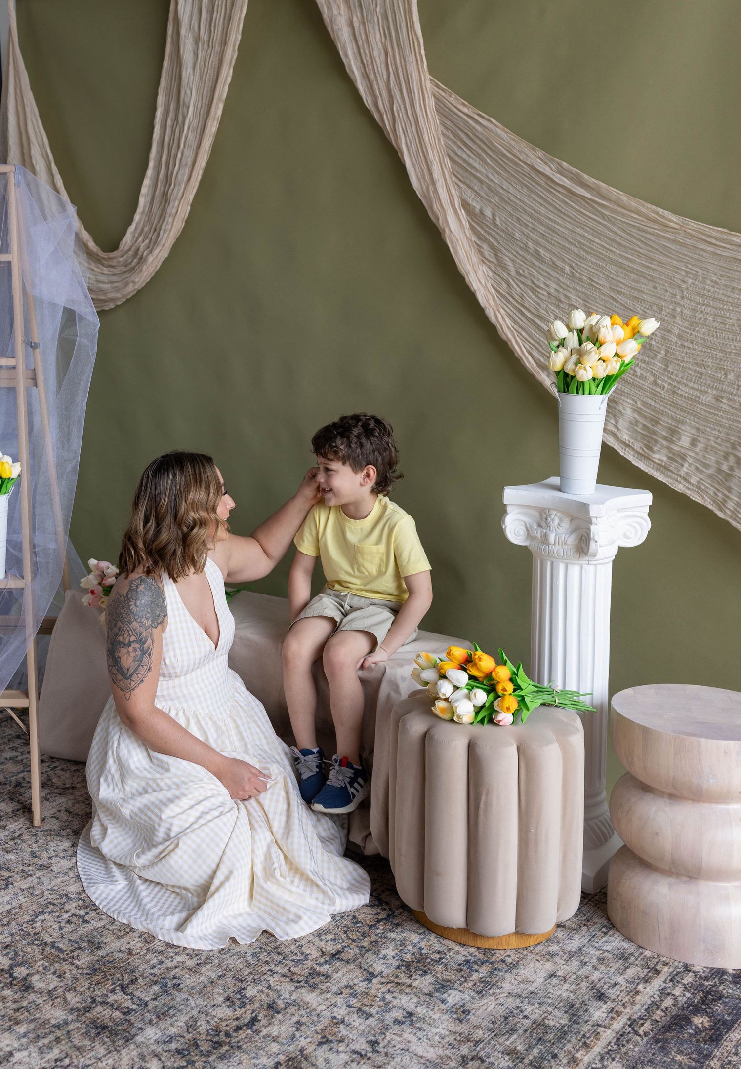 A mother in a white dress sits with her young son in a yellow shirt in a green studio with floral decor.