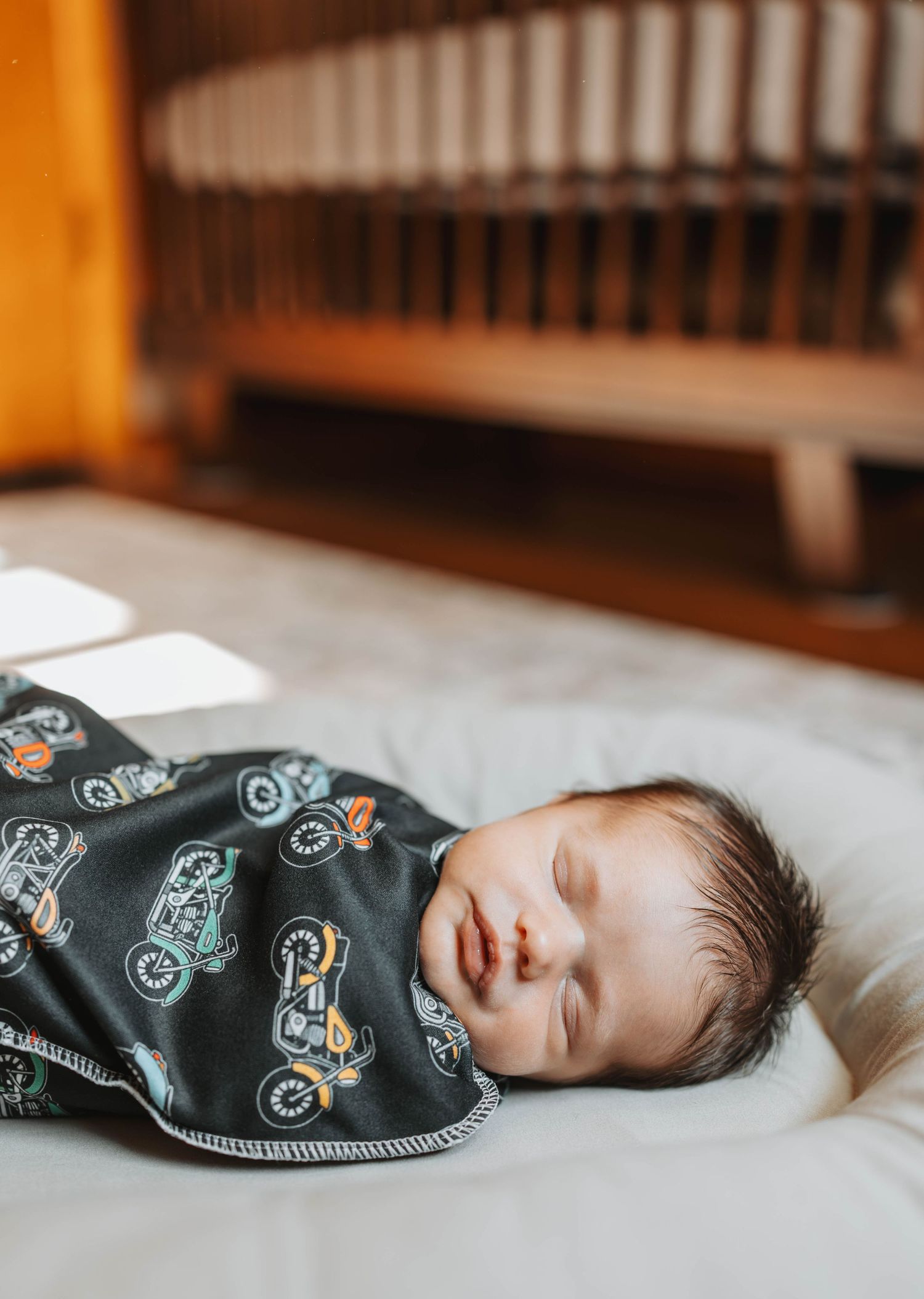 Sleeping newborn baby swaddled in a dark motorcycle print blanket lying on a white mattress near a wooden crib.