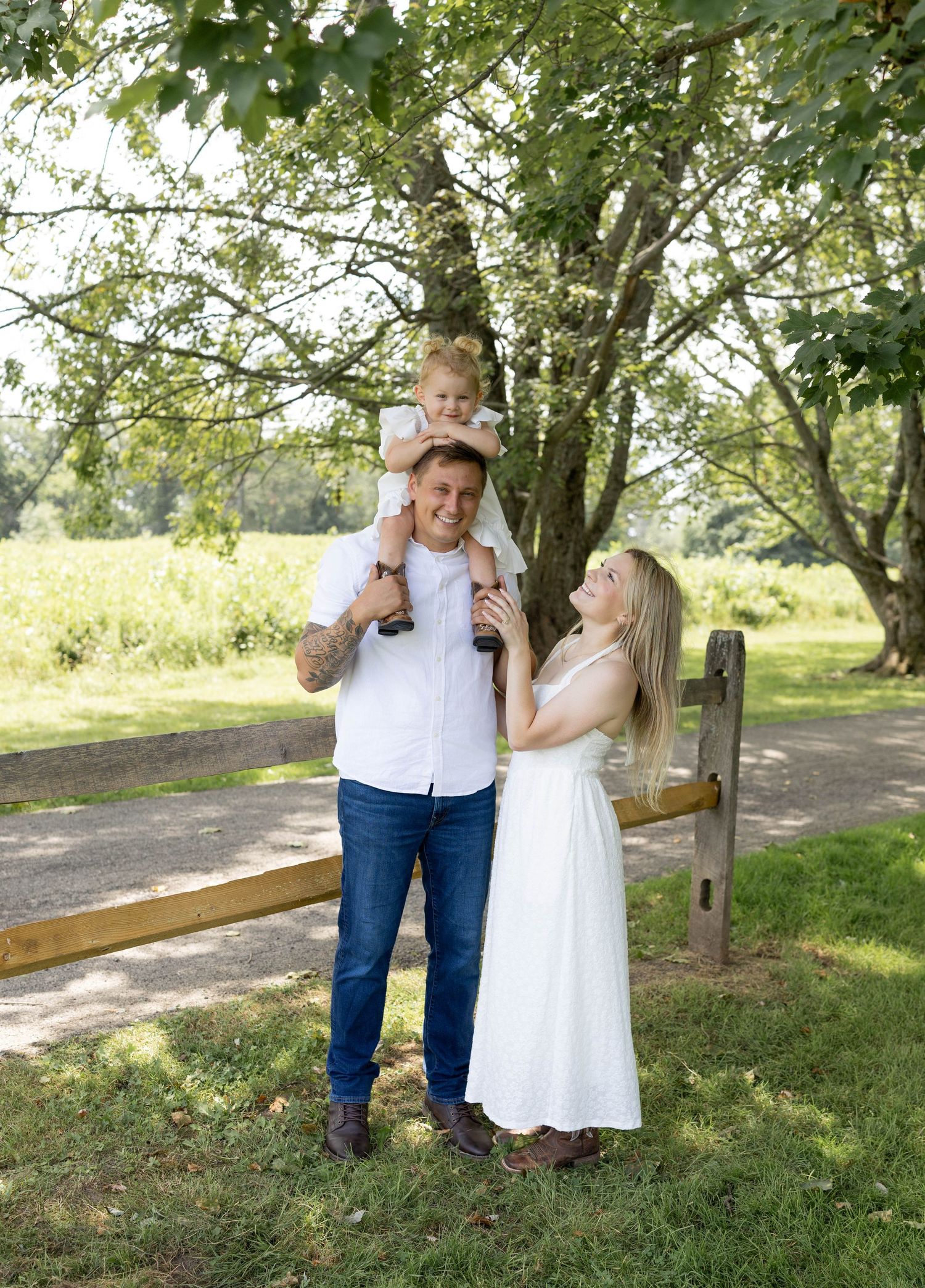 Family of three posing outdoors, father carrying toddler on shoulders beside mother in white dress near wooden fence.
