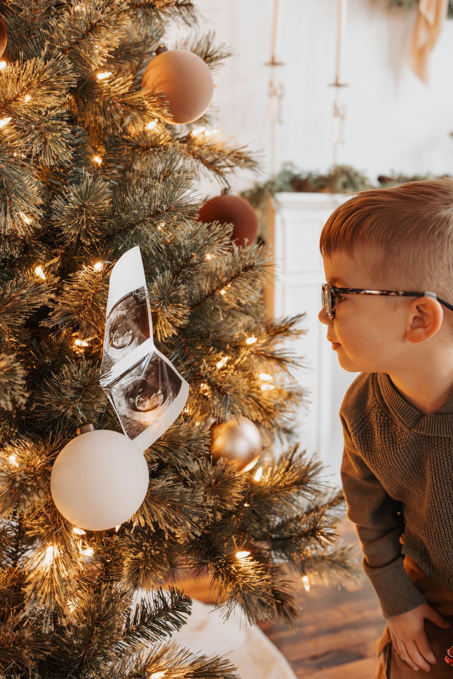 A white Christmas ornament hangs on an illuminated evergreen tree branch with warm glowing lights in the background.