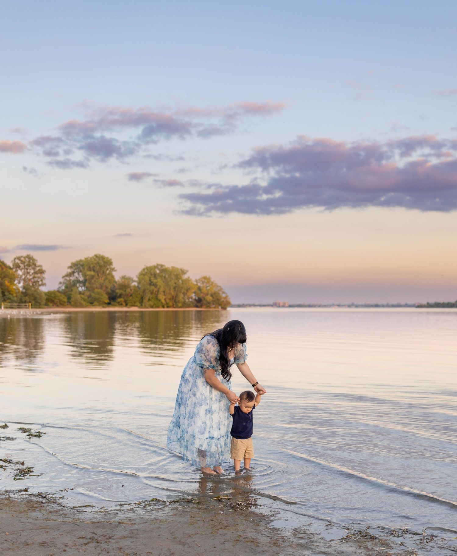 Two figures stand in shallow water at sunset by a tranquil lake shore with trees silhouetted against a pastel sky.