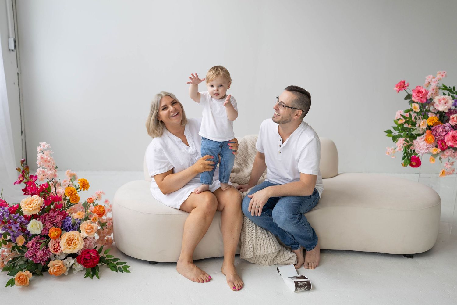 Three people sit together on a beige couch with bright colorful floral arrangements on white background.