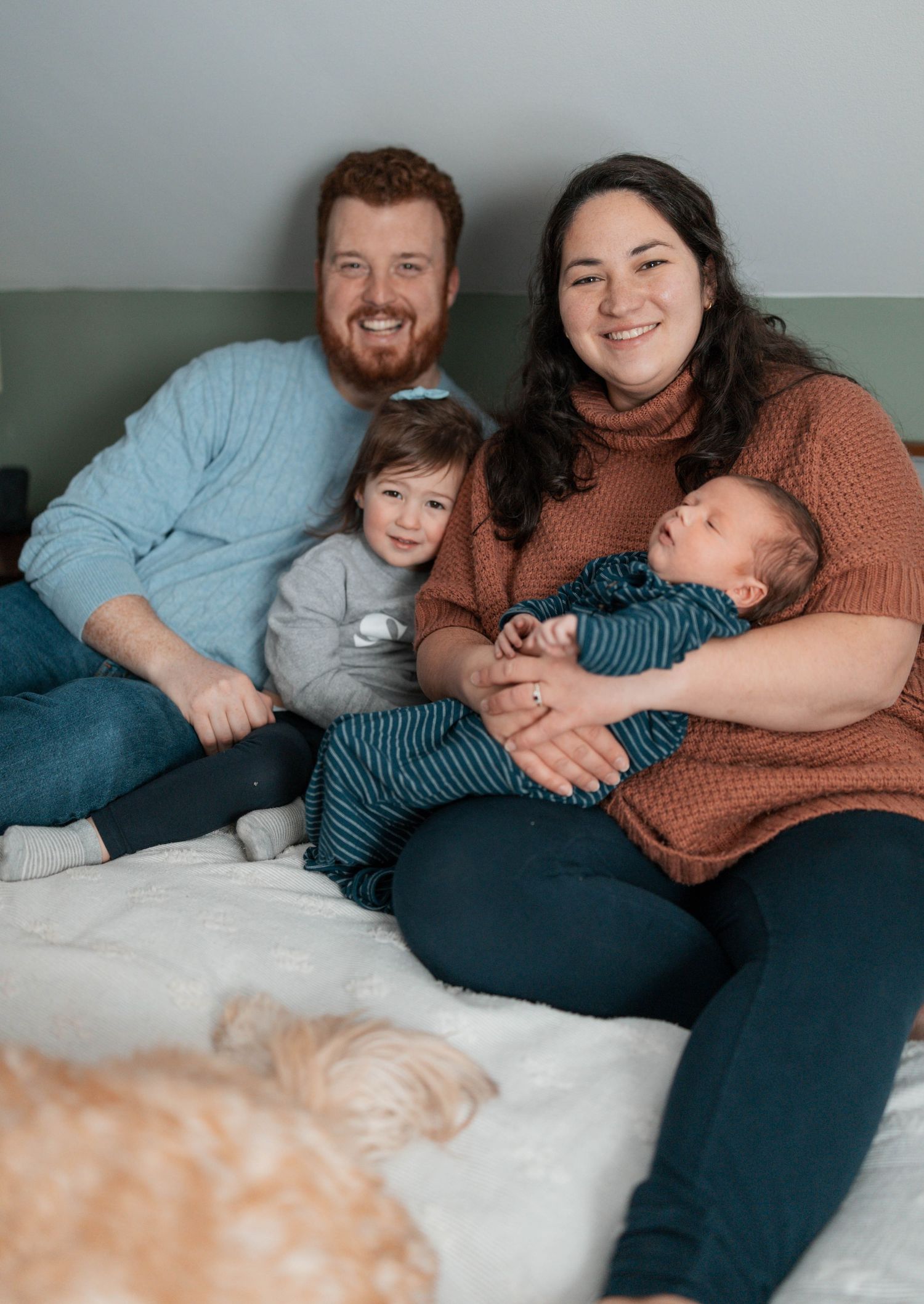 A casual family portrait on a couch showing parents with two young children and a glimpse of their pet dog.