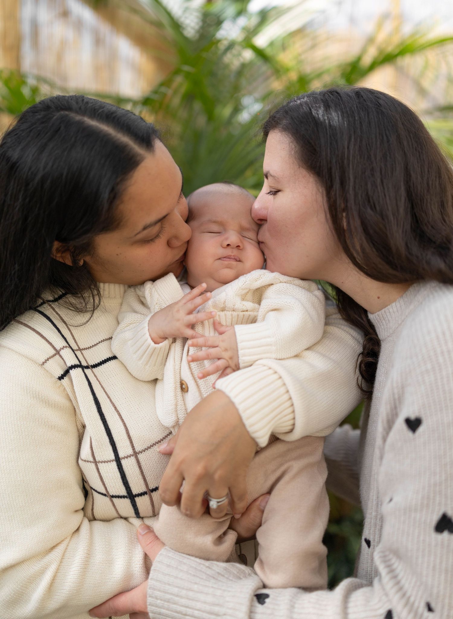 Two adults lovingly kiss a sleeping baby in soft knit sweaters surrounded by greenery.