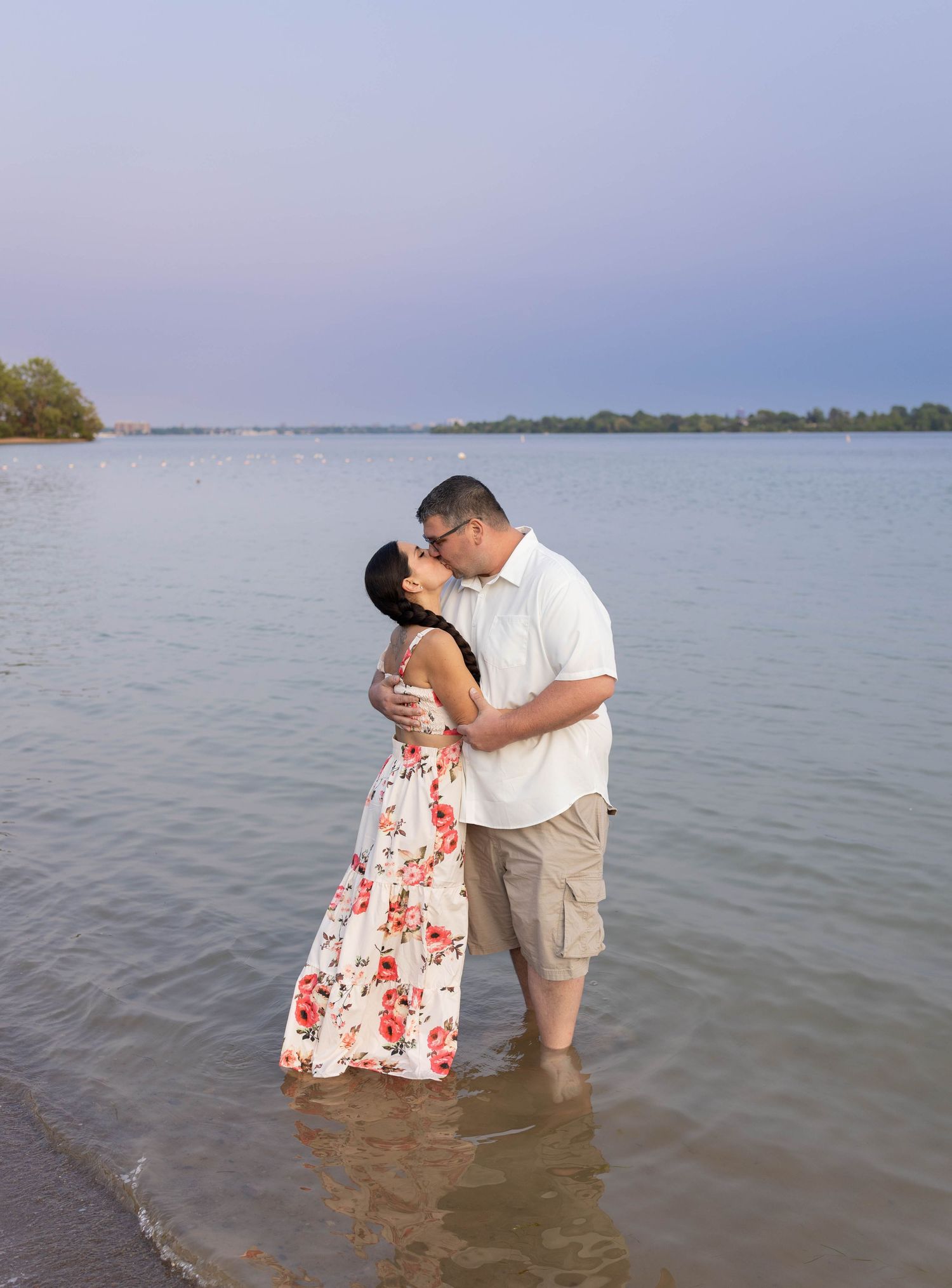 Couple kissing while standing in shallow lake water, woman in floral dress, man in white shirt and khaki shorts.