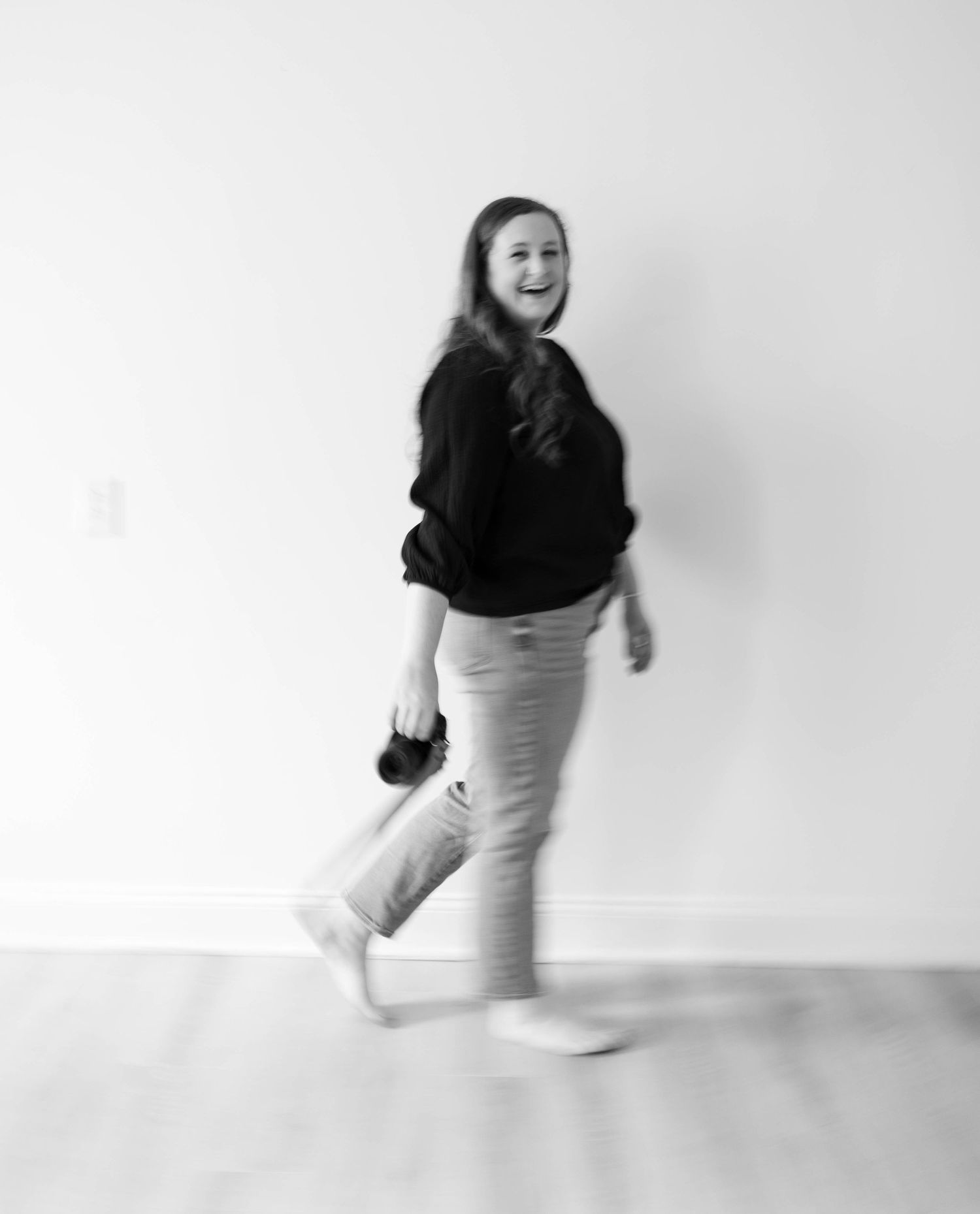 Black and white photo of someone jumping playfully in casual attire against a white studio backdrop.