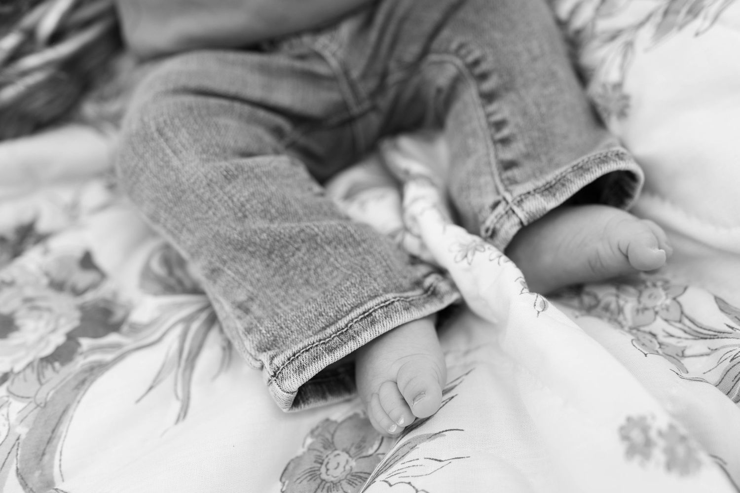 Black and white closeup of tiny toes peeking out from the cuff of faded blue jeans on floral bedding.