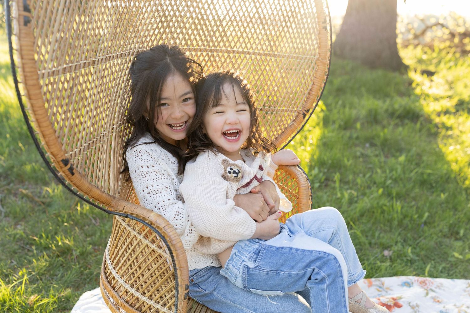 Two people sitting together in a peacock chair on grass share joyful laughter while embracing outdoors in sunlight.