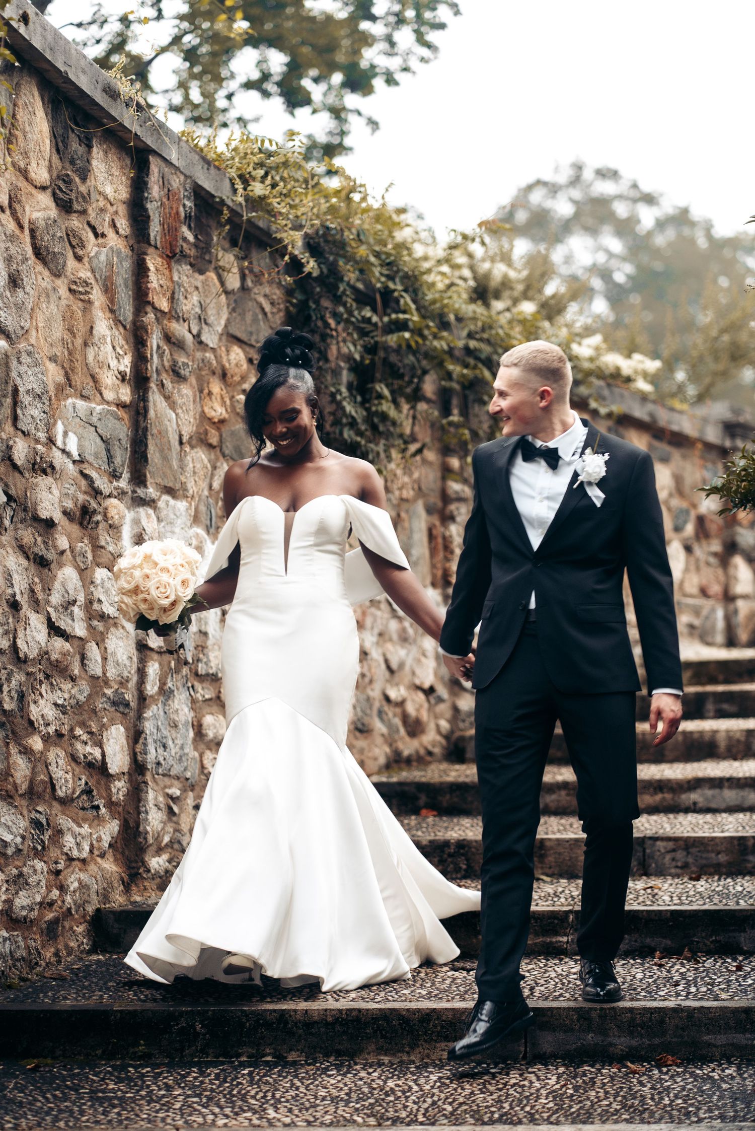 A bride and groom descend stone steps on their wedding day, with a rustic stone wall and greenery backdrop.