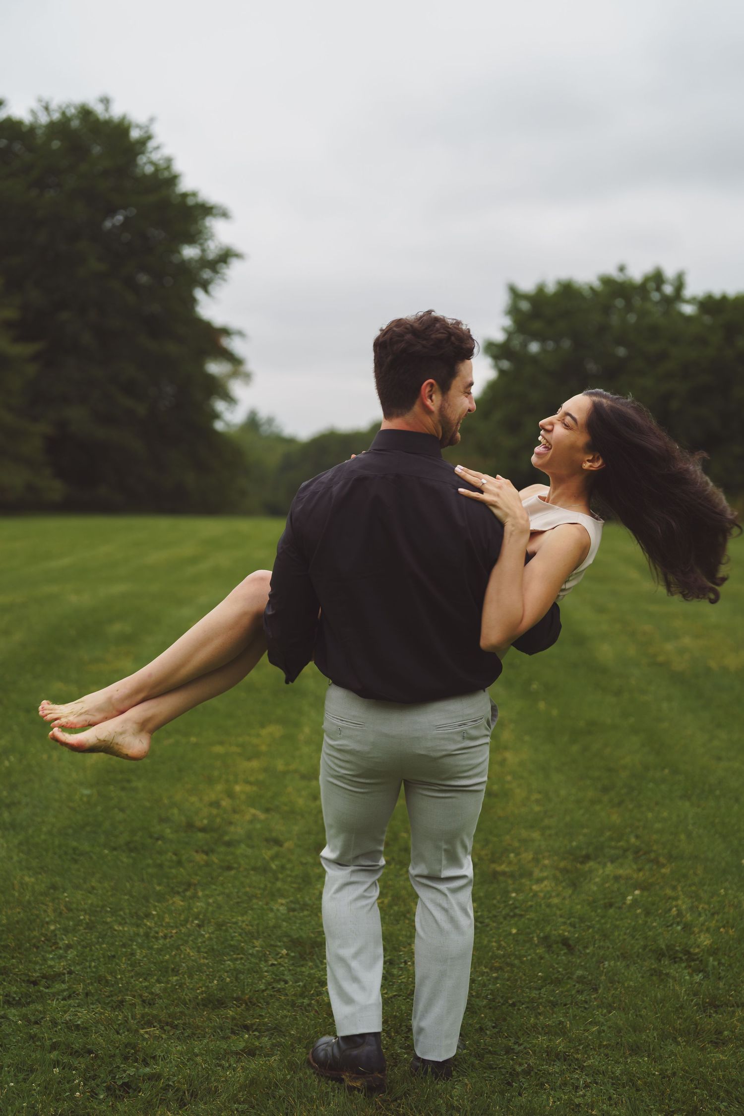 A romantic couple shares a joyful moment in a grassy field as one person carries the other against an overcast sky.