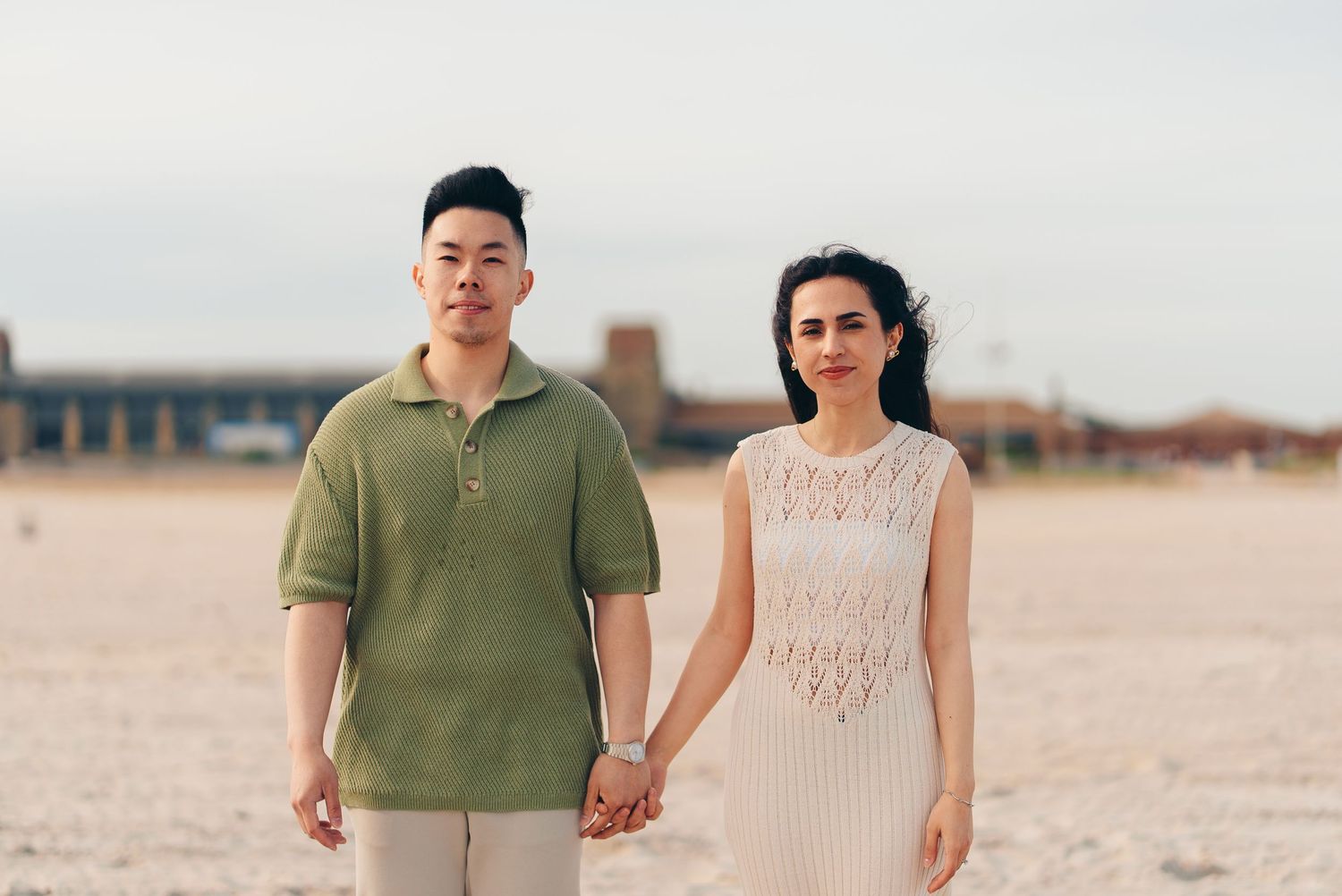 A couple holding hands on a beach wearing a green polo shirt and cream knit dress respectively.