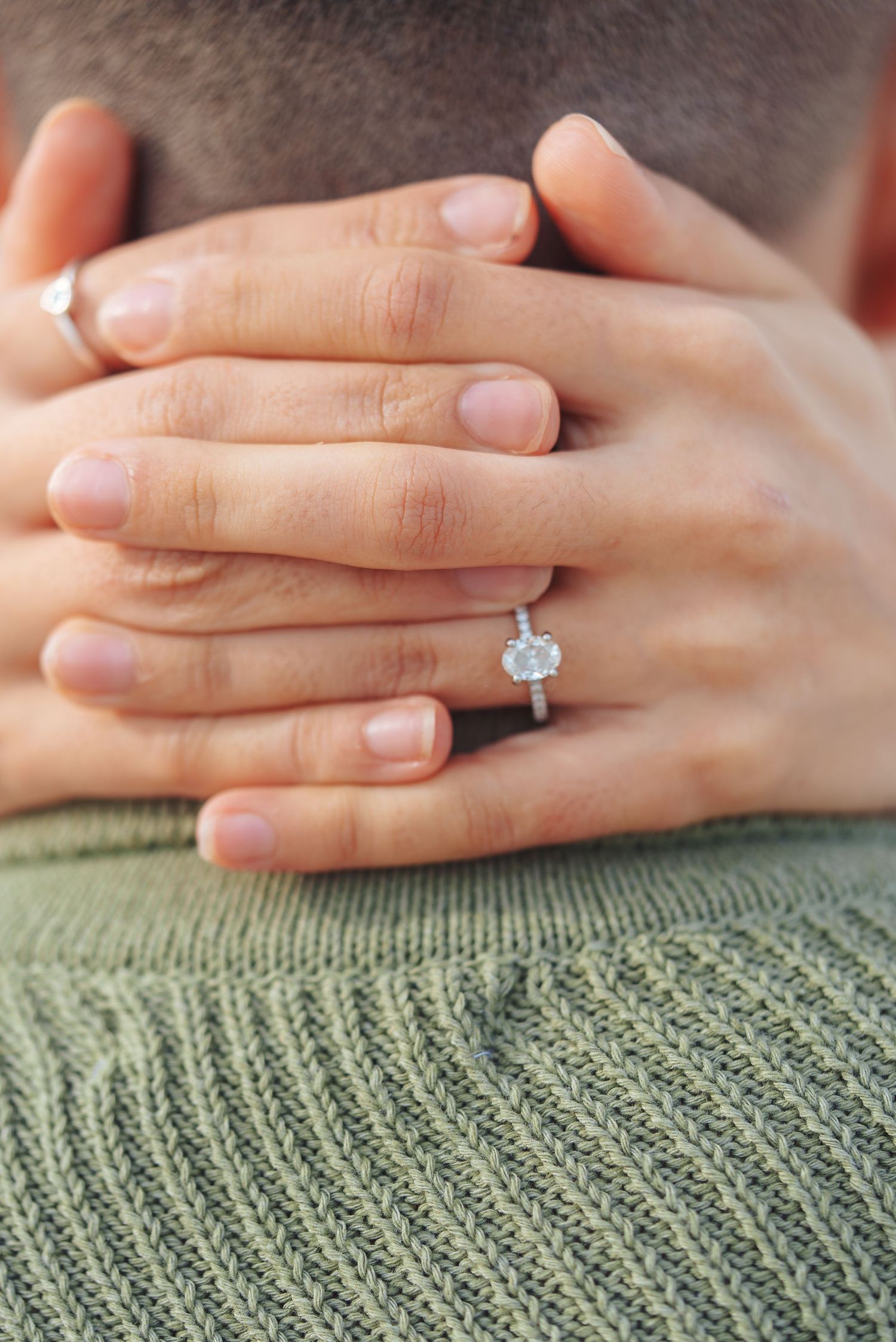 Close-up of hands with a diamond engagement ring against a green knit sweater backdrop.
