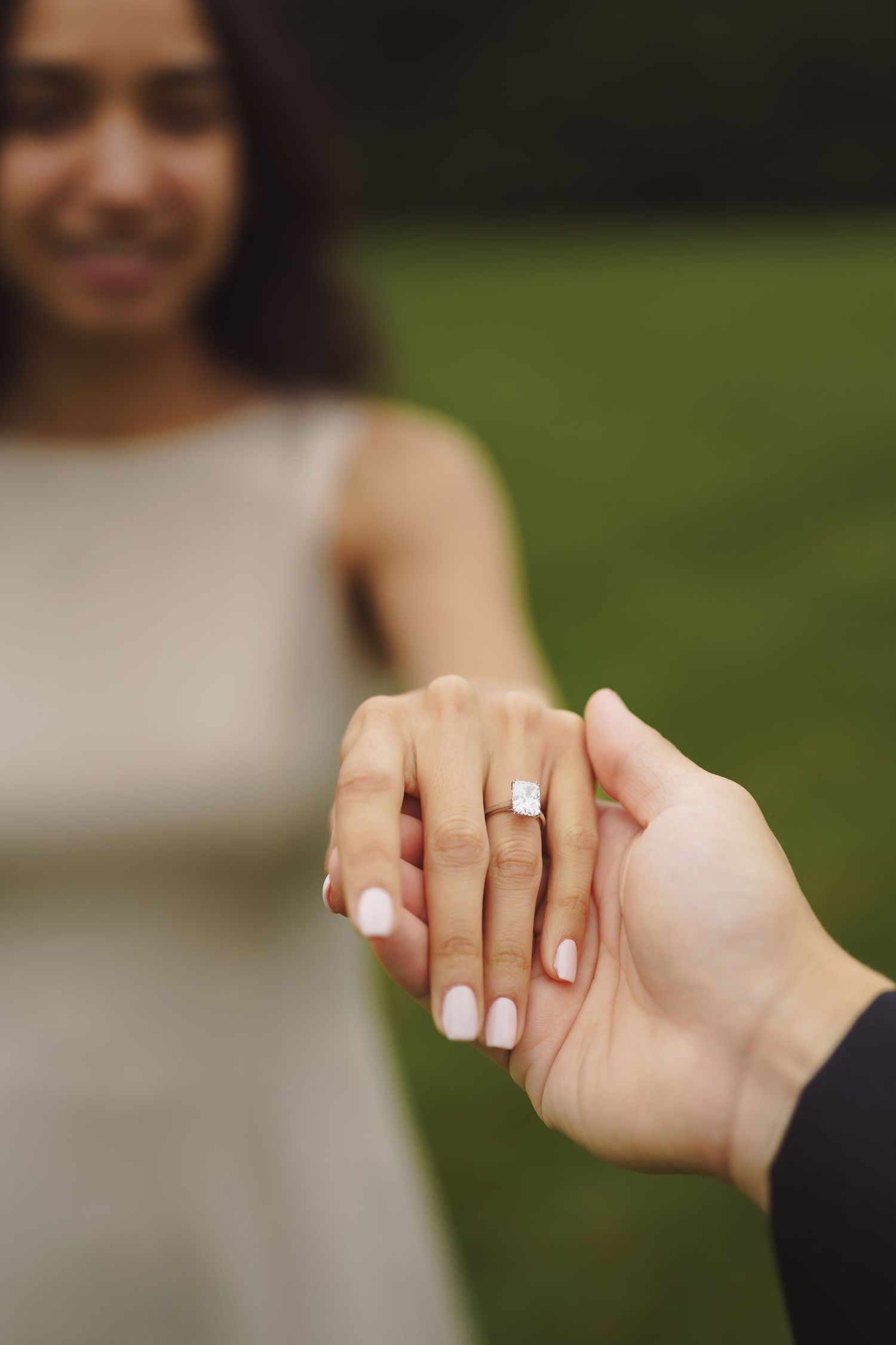Two hands intertwined with an engagement ring sparkling in focus against a blurred green outdoor background.