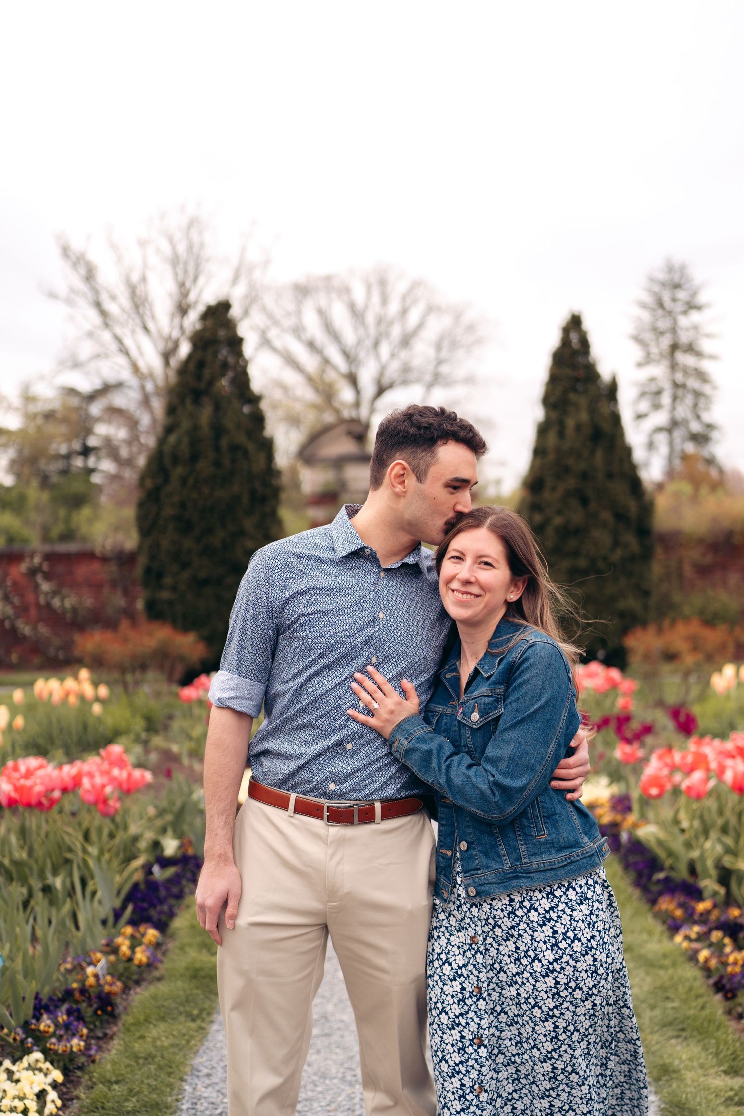 A couple shares a tender moment while standing in a garden filled with blooming tulips and evergreen shrubs.