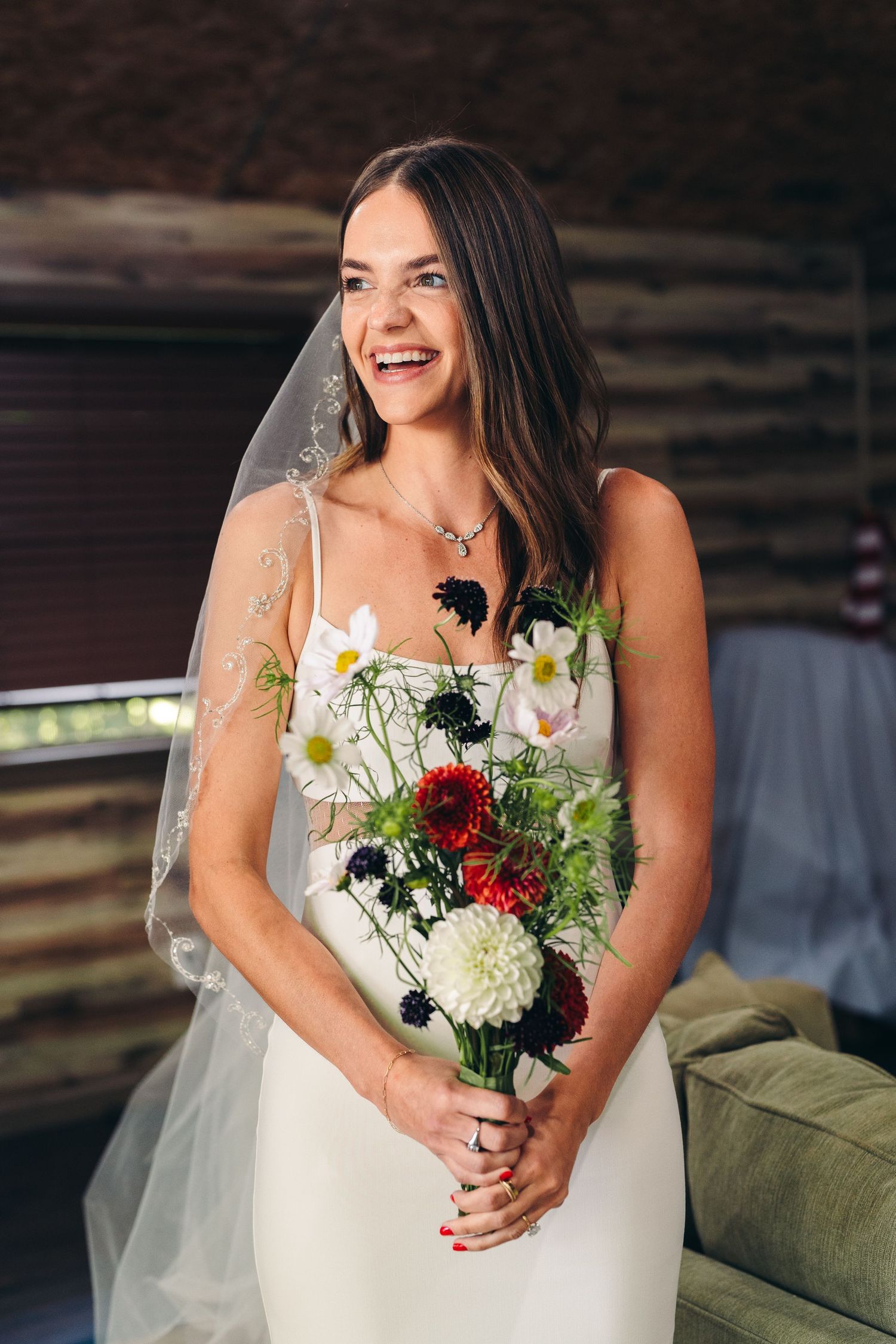 A bride in a white wedding dress laughs while holding a colorful bouquet with white, red and green flowers.