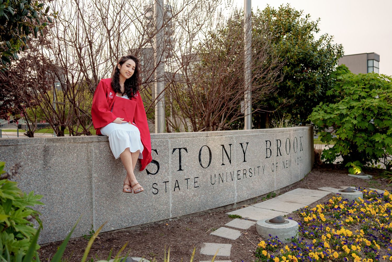 Student in red sweater sits on Stony Brook University entrance sign surrounded by yellow flowers and greenery.