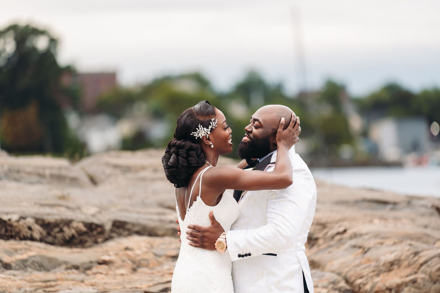 A happy couple shares a romantic embrace on a beach while dressed in elegant white wedding attire.