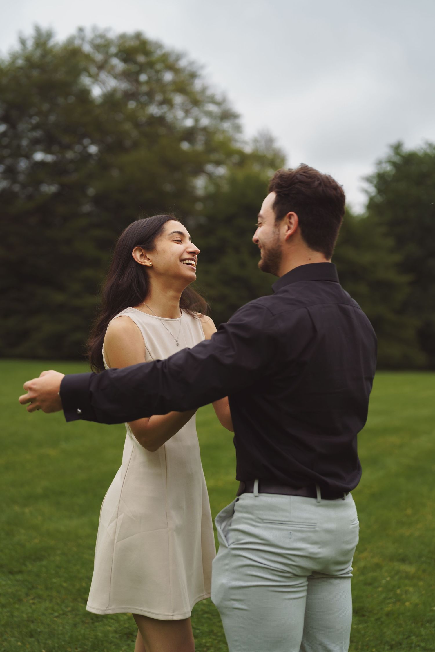A couple shares a joyful moment dancing together on a grass lawn surrounded by trees on an overcast day.