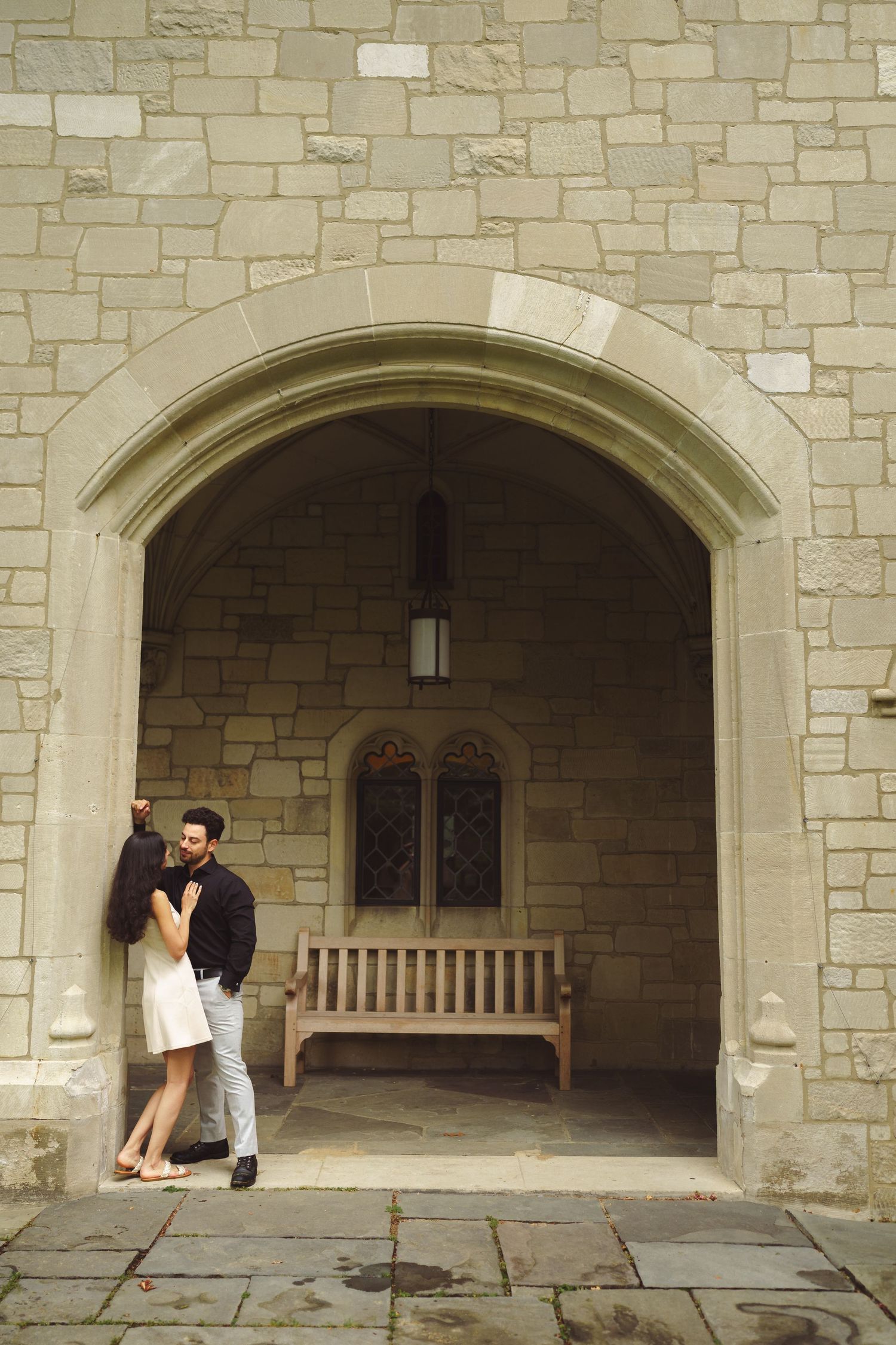 A couple embraces in a stone archway alcove with a wooden bench and gothic architectural details.