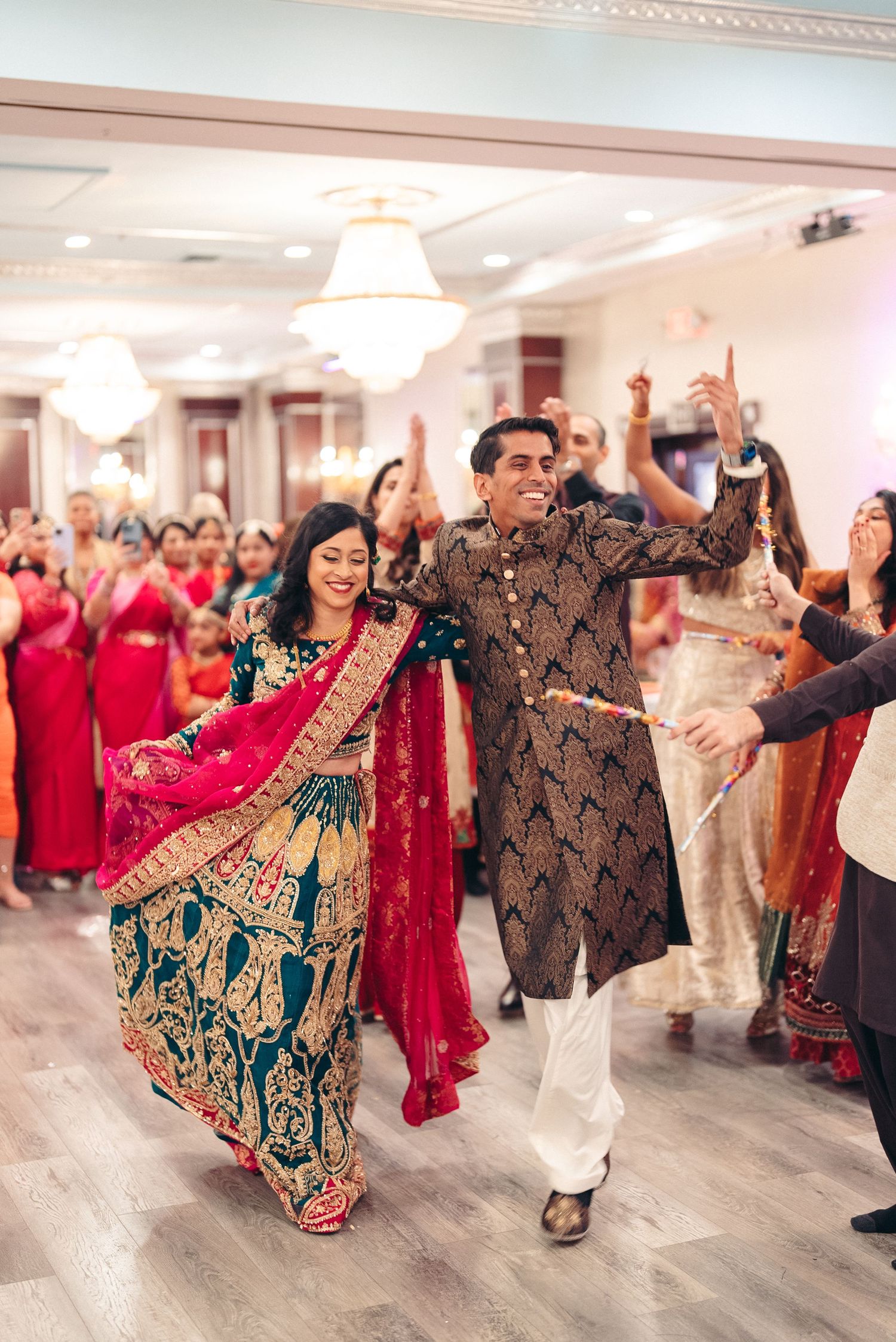 Festive dancers in traditional Indian wedding attire celebrate on dance floor during reception with guests in red outfits.