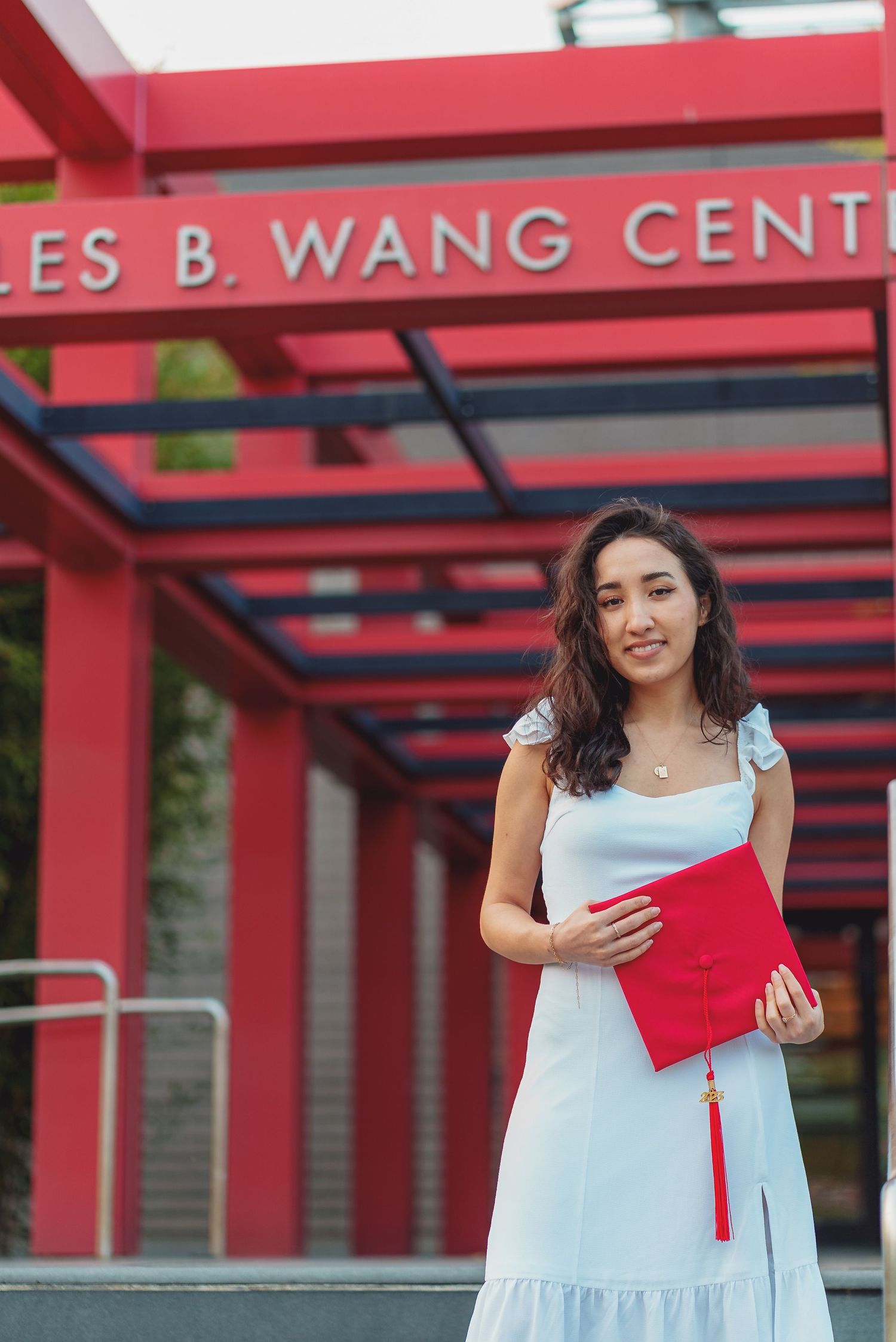 Graduate in white dress holds red diploma folder while standing under the Charles B. Wang Center sign.