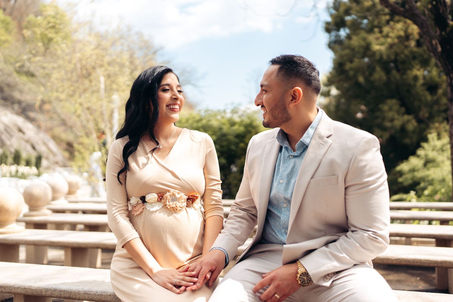 A couple sits together on concrete steps outdoors holding hands and smiling at each other on a sunny day.