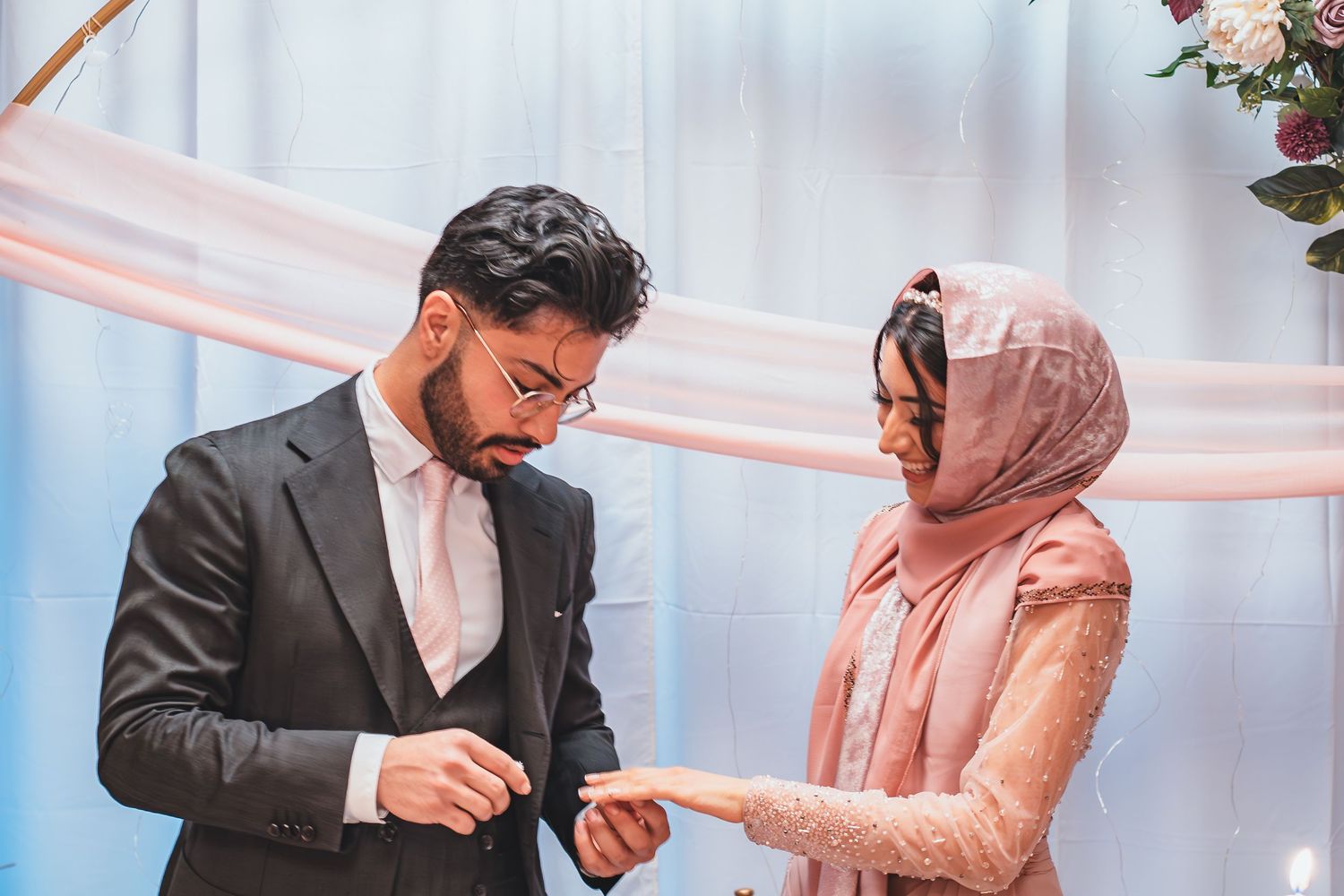 A couple in formal attire exchanges rings during their ceremony against a decorated blue backdrop.