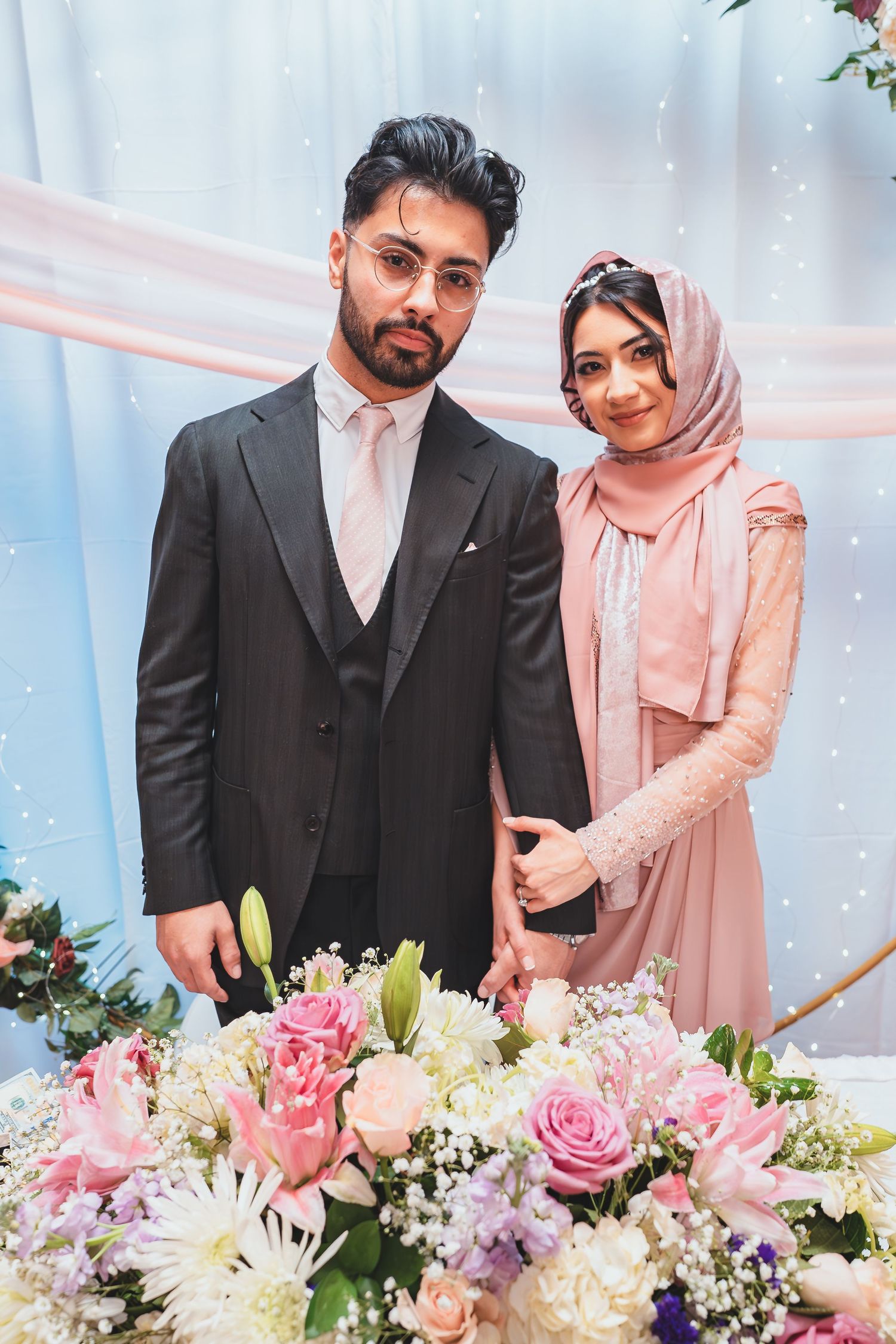 A formal couple poses by a floral arrangement with pink roses and white lilies against a light blue backdrop.