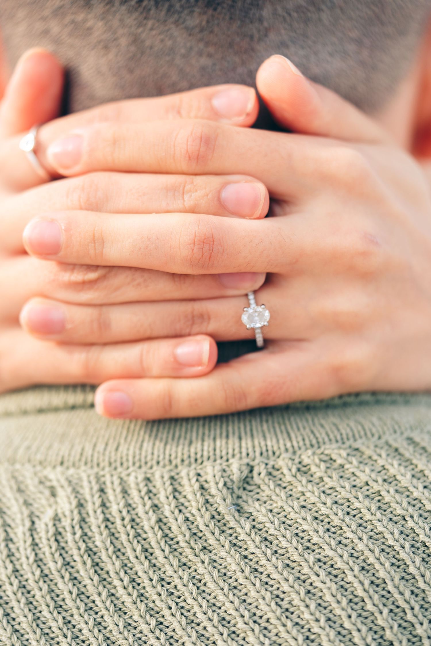 Close-up of hands with a diamond engagement ring resting on a gray knit sweater.