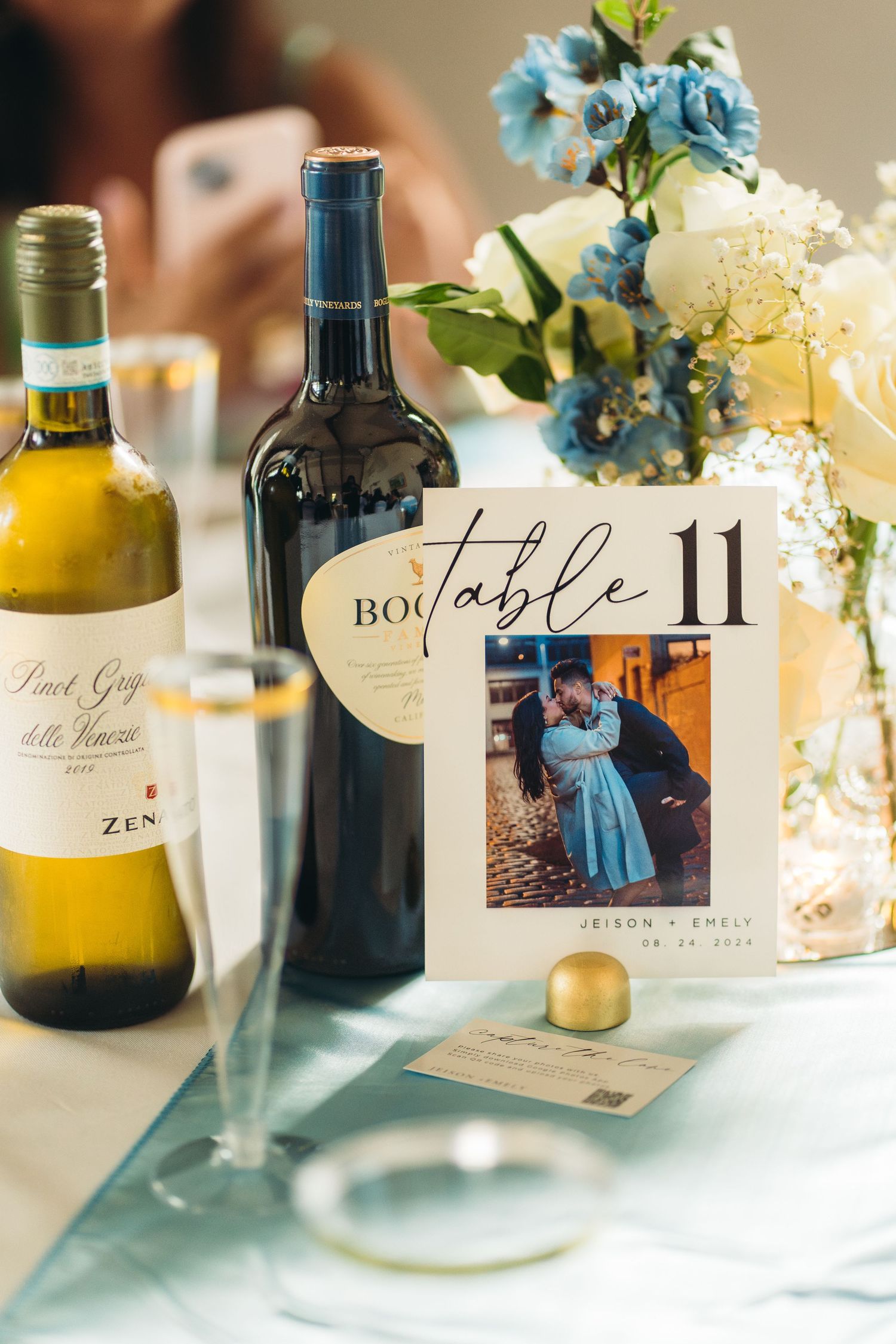 Wedding table setting with wine bottles, floral centerpiece and vintage-style table number display featuring a photograph.