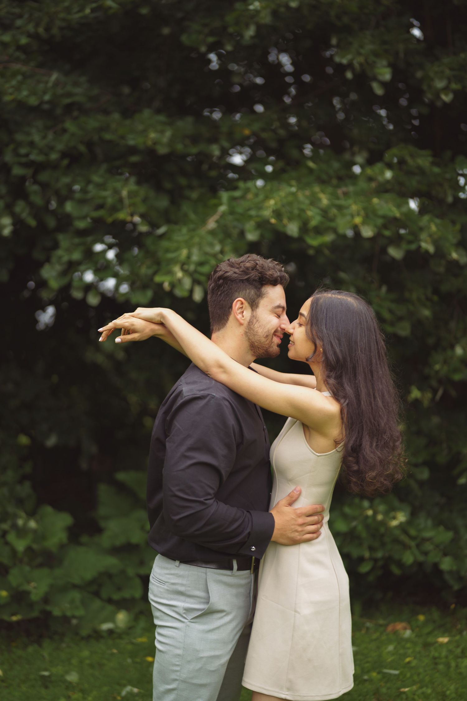 A romantic couple embraces and kisses in a lush garden setting with green foliage in the background.