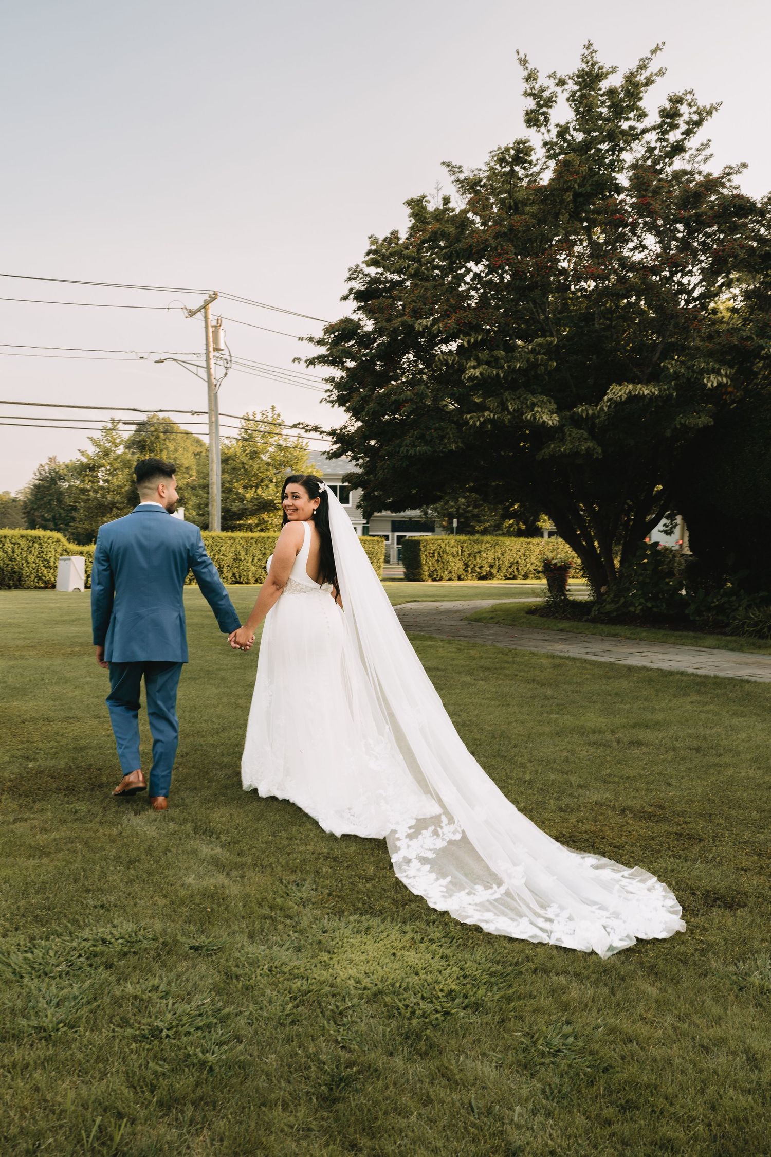 A wedding couple walks hand in hand on a grassy lawn with a flowing white veil trailing behind the bride.