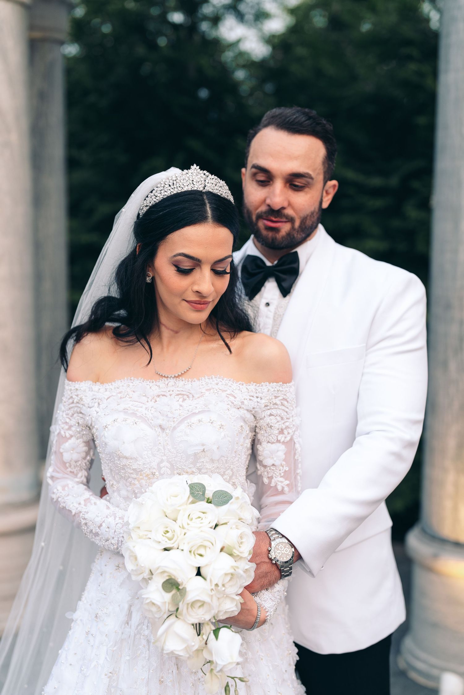 Elegant couple in formal wedding attire with white floral bouquet posing outdoors by classical columns.
