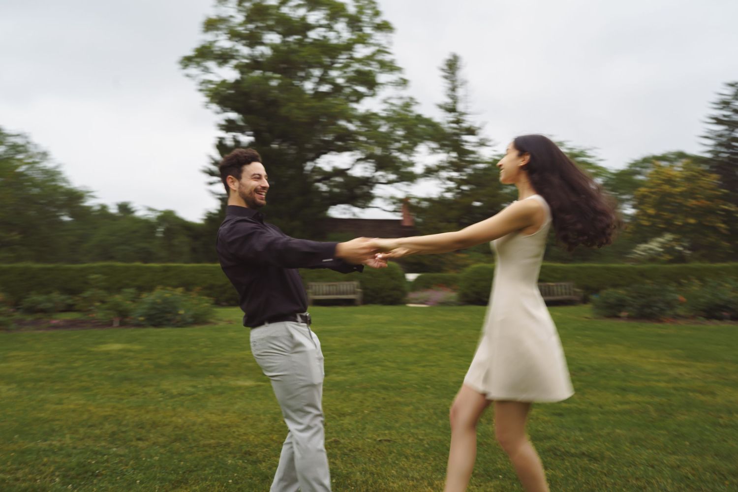 Two people hold hands while spinning and dancing together on a green lawn surrounded by trees.