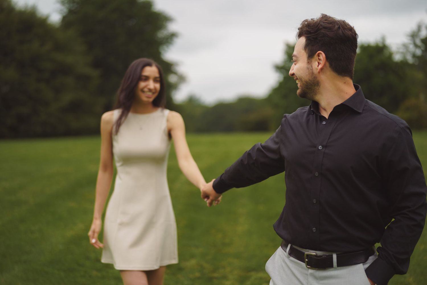 A couple holds hands while walking together on a grassy field under an overcast sky.