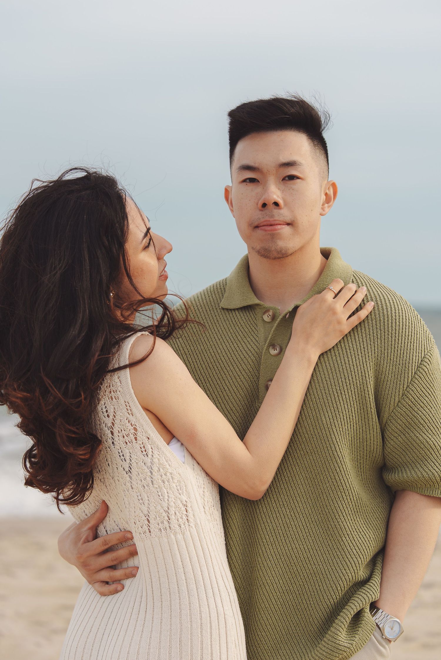 Romantic couple embraces on beach, wearing olive green polo and white lace dress against cloudy sky backdrop.