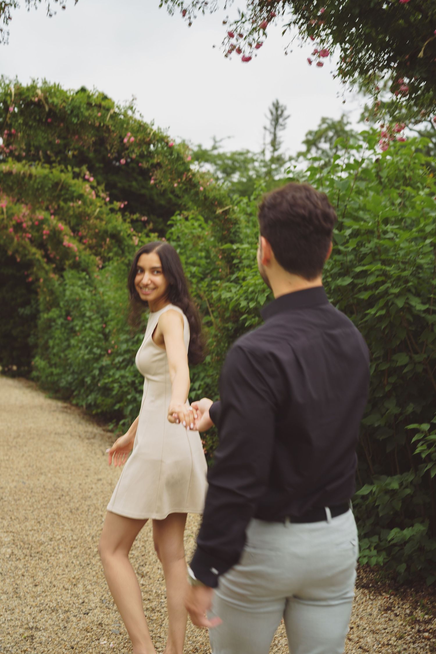 A couple dances and twirls together along a garden path lined with flowering bushes.