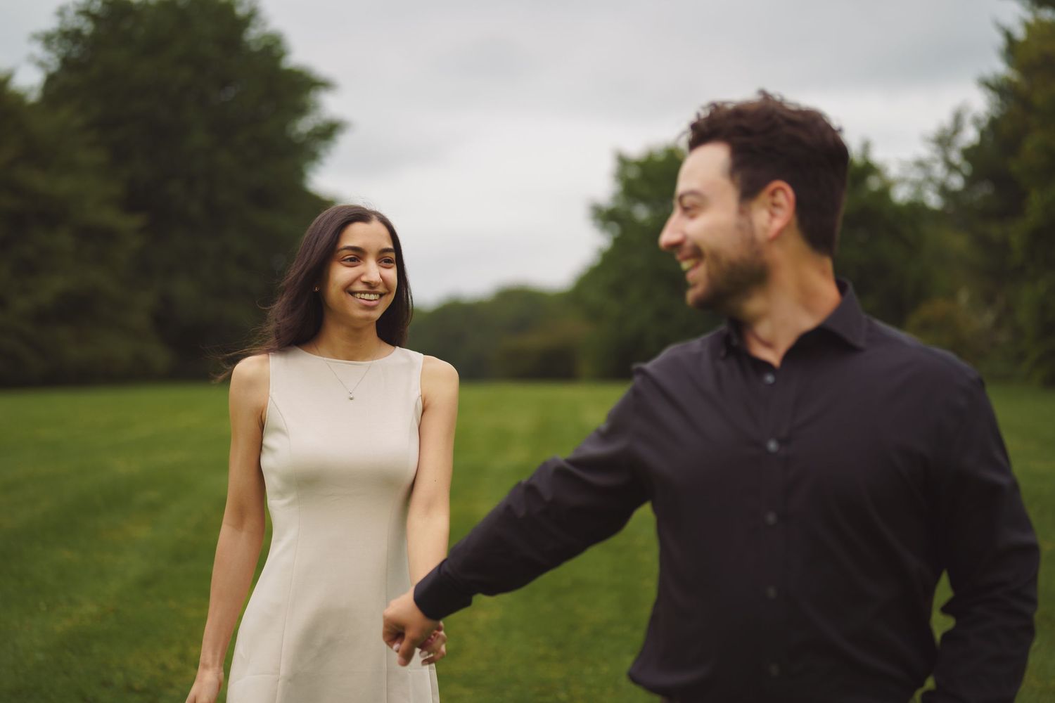 A couple in wedding attire smiles and laughs together while walking outdoors in a green field on their special day.
