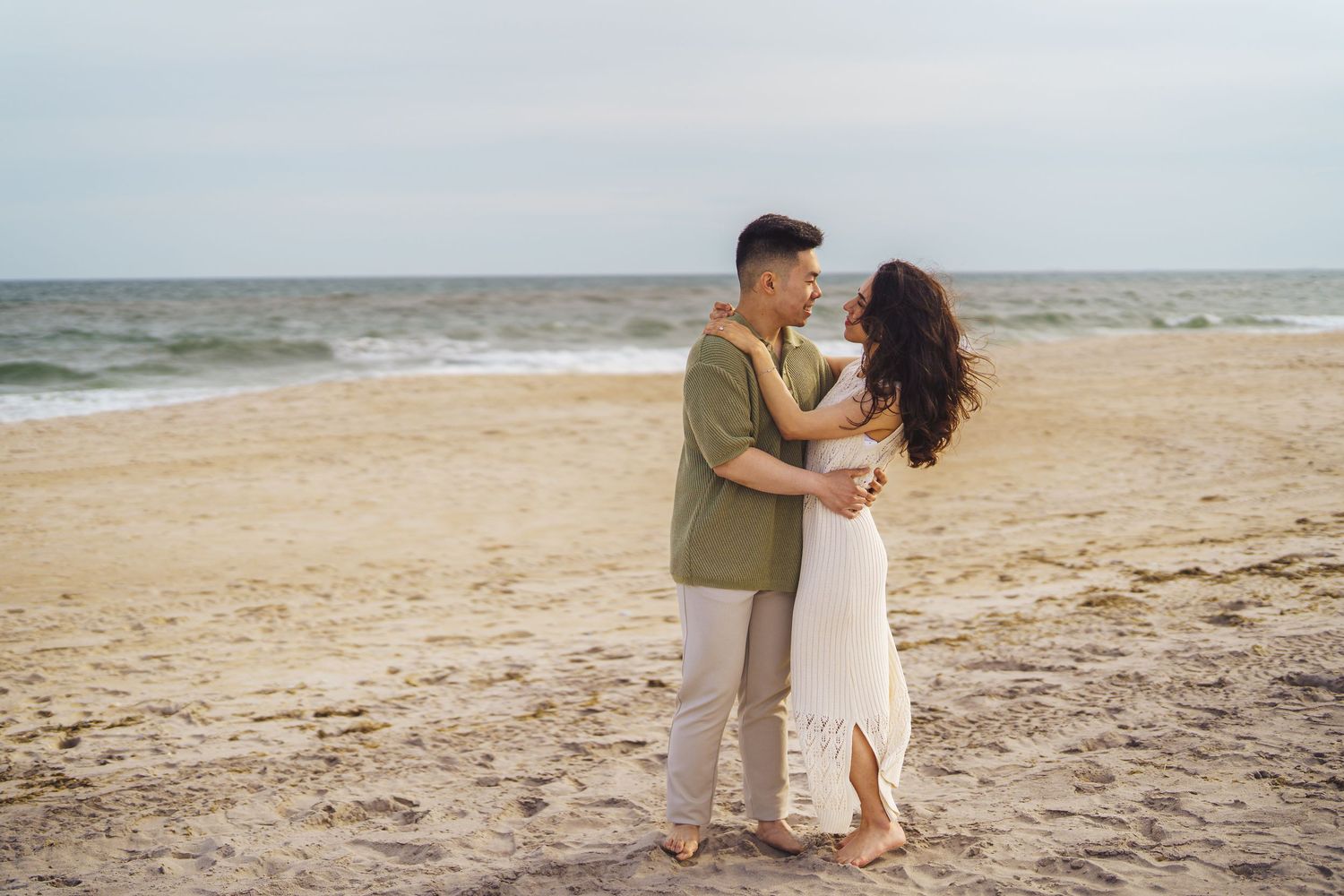 A couple embraces tenderly on a sandy beach with ocean waves rolling in behind them on a cloudy day.